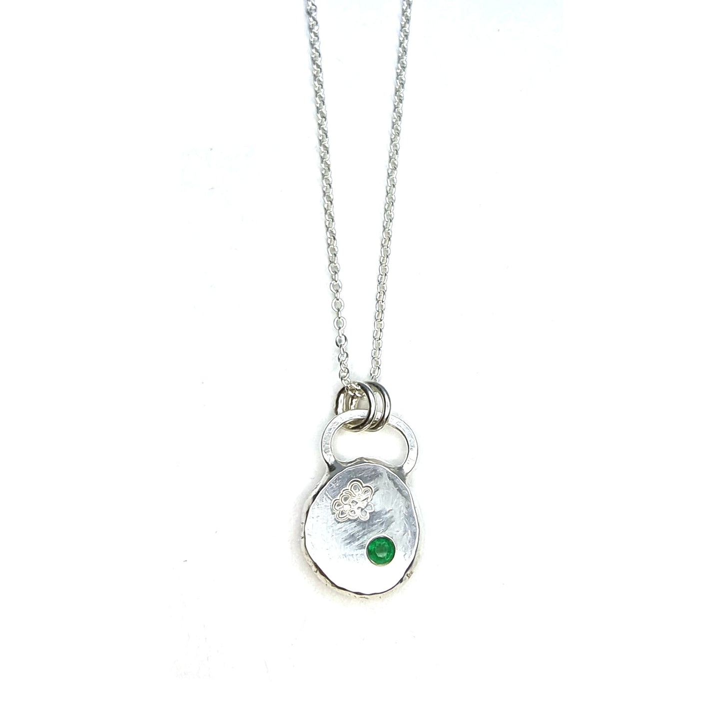 Round silver pendant necklace with a hawthorn flower engraved on it and a flush set green emerald gemstone. On a silver chain.