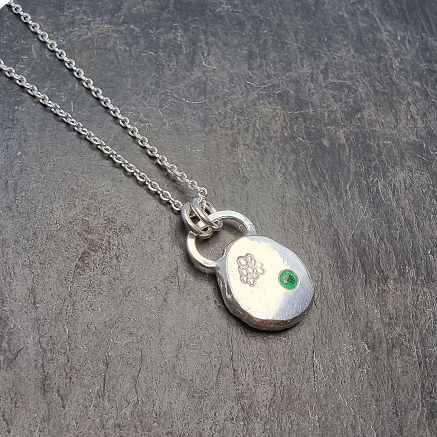 Round silver pendant necklace with a hawthorn flower engraved on it and a flush set green emerald gemstone. On a silver chain.