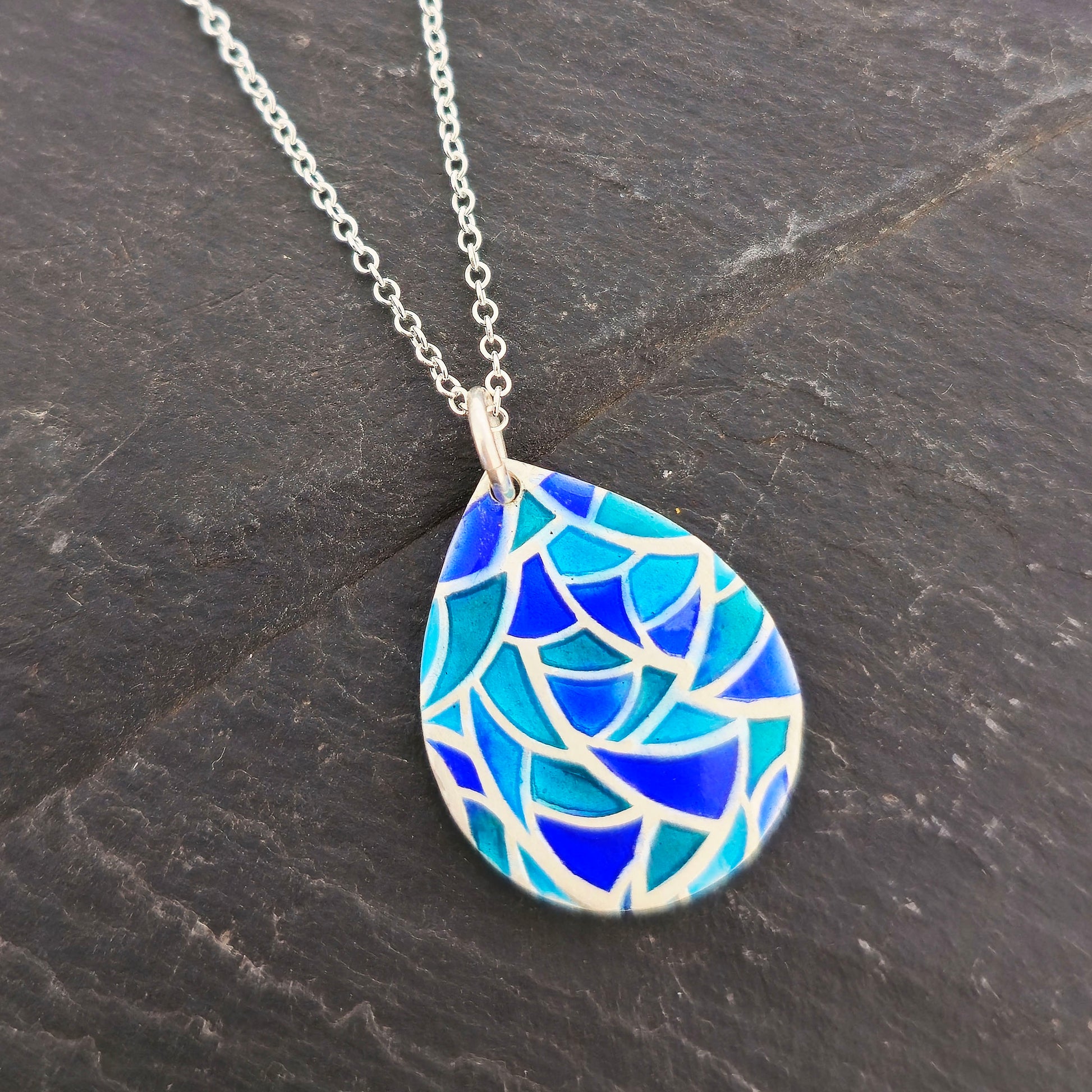 A teardrop shaped silver pendant with geometric cells of blue, green and turquoise enamel. On a silver chain. Pictured on a black background. Mijoux.