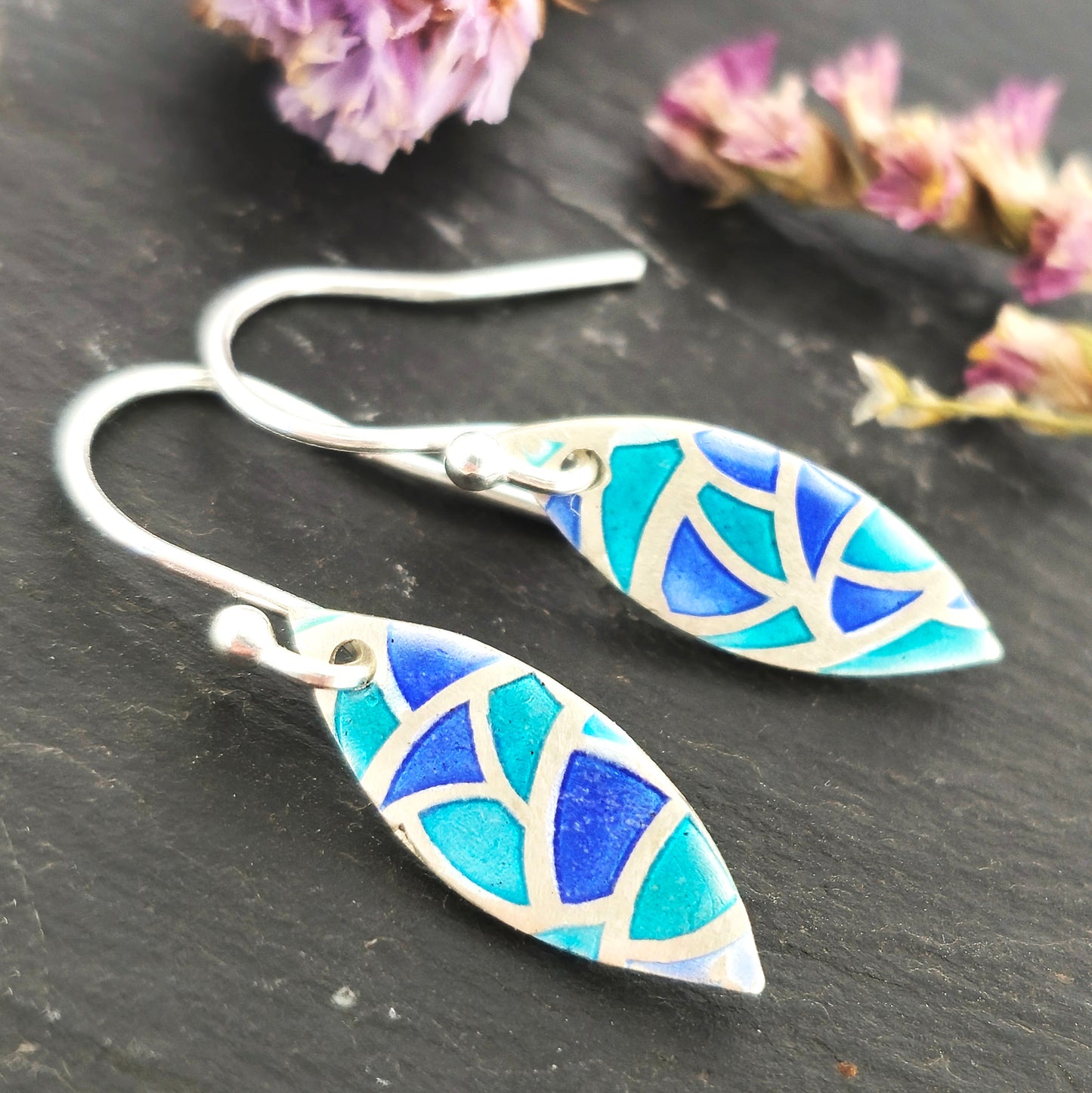 Silver drop earrings. Each earrings has a marquise shape with geometric cells containing blue, green and turquoise enamel. Pictured on a black surface with dried flowers.