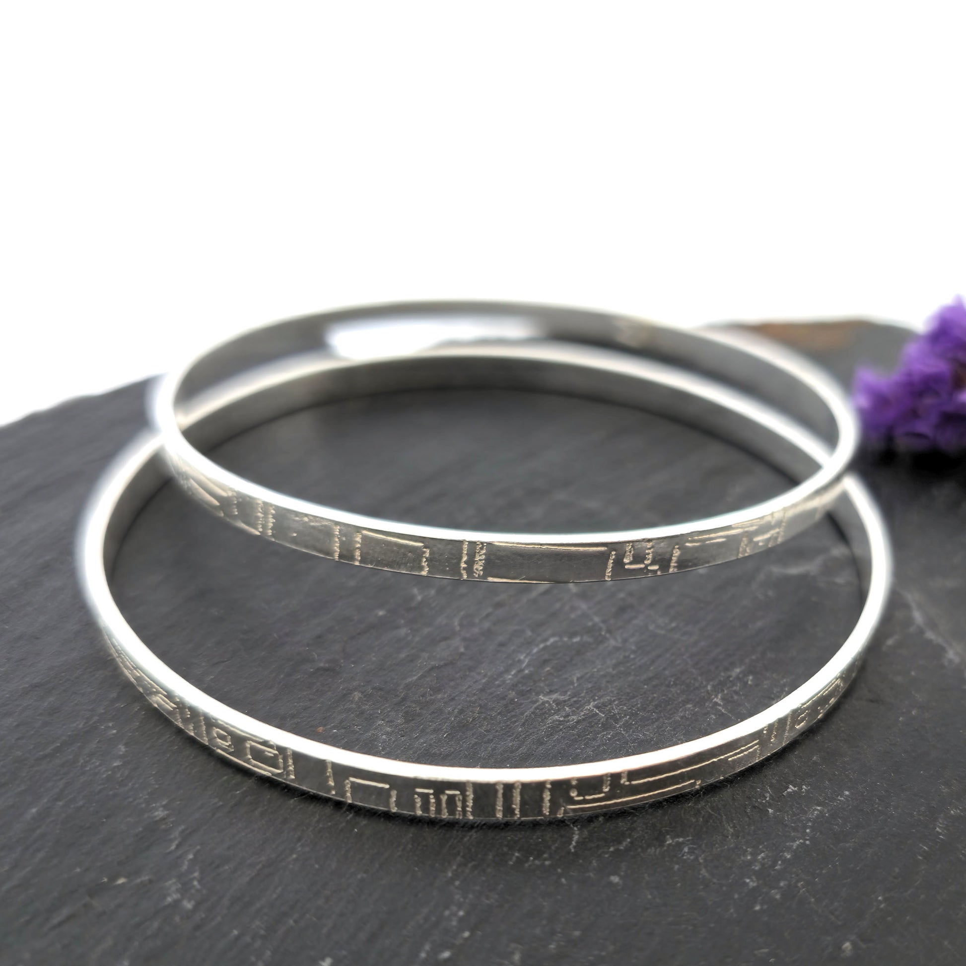 Two flat silver bangles with a square and line pattern on a dark surface with a purple flower. Mijoux Jewellery.