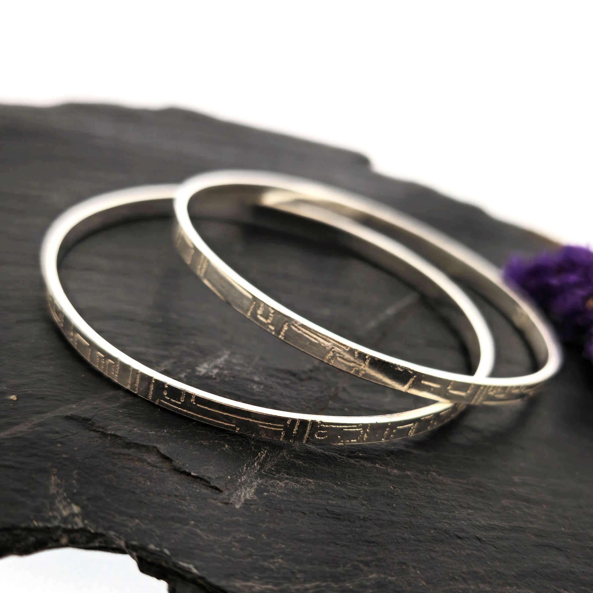 Two flat silver bangles with a square and line pattern on a dark surface with a purple flower. Mijoux Jewellery.
