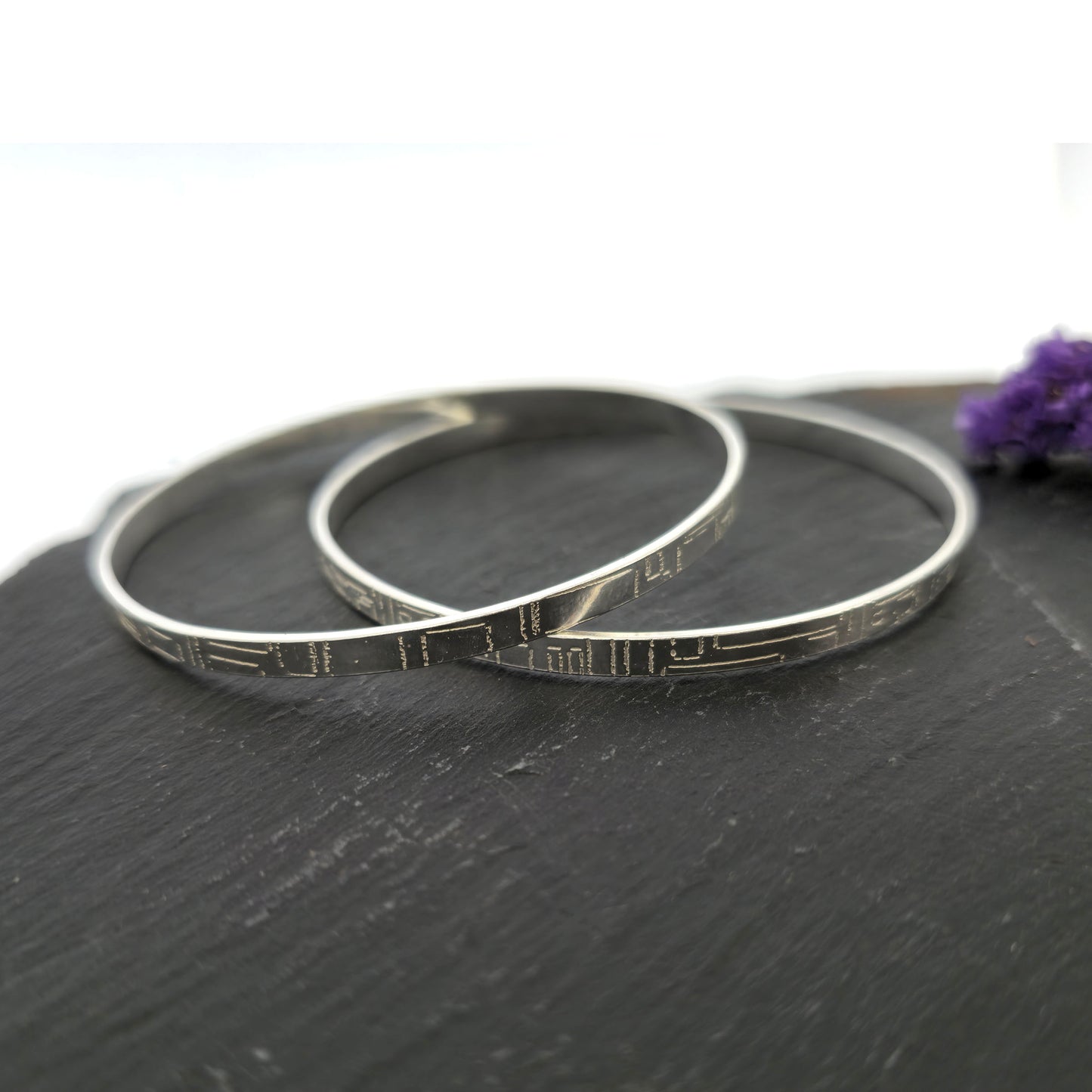 Two flat silver bangles with a square and line pattern on a dark surface with a purple flower. Mijoux Jewellery.