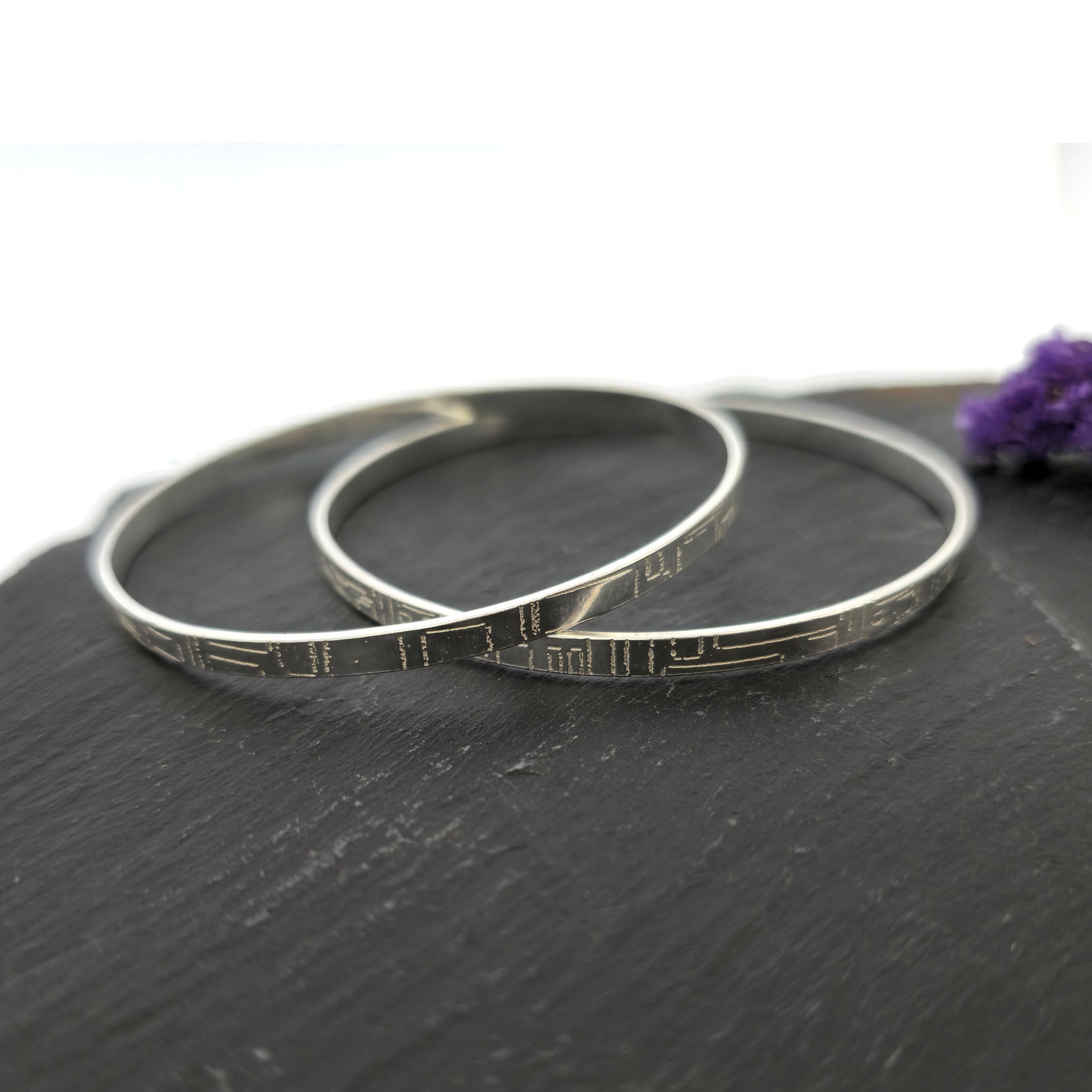 Two flat silver bangles with a square and line pattern on a dark surface with a purple flower. Mijoux Jewellery.
