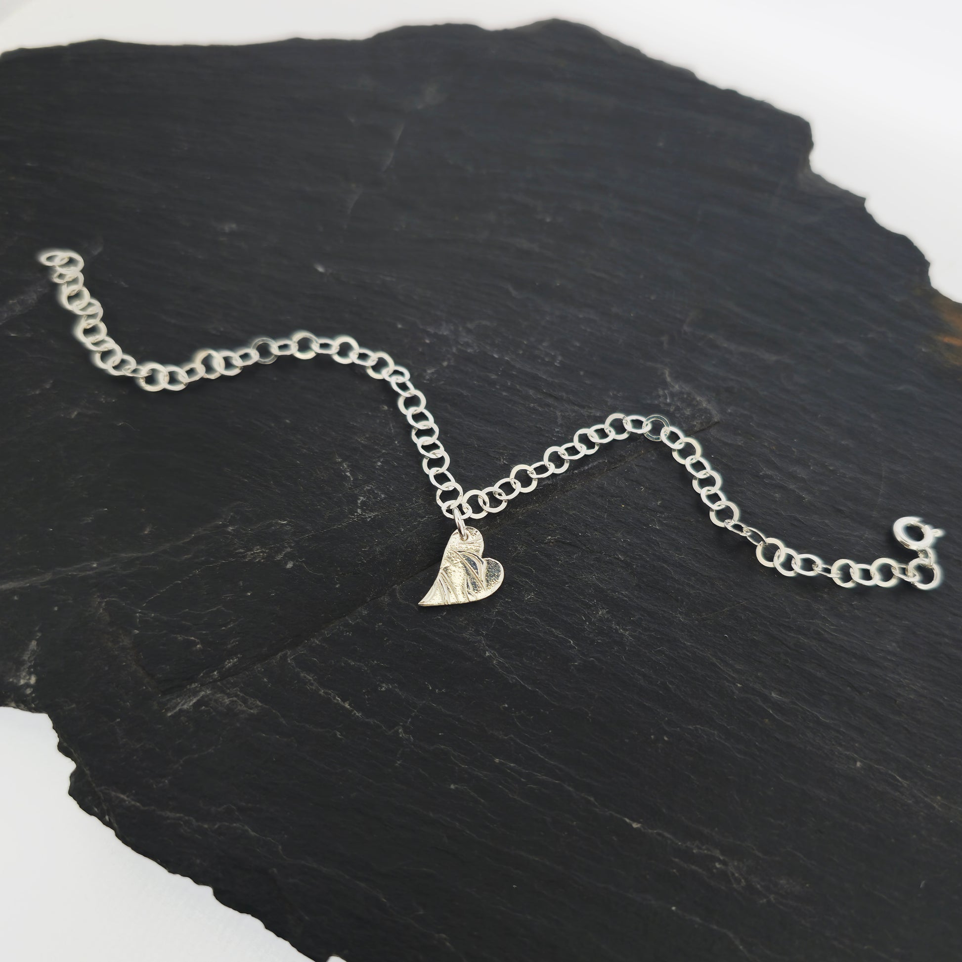 Silver large link chain bracelet with asymmetrical patterned heart charm. Shown on slate.