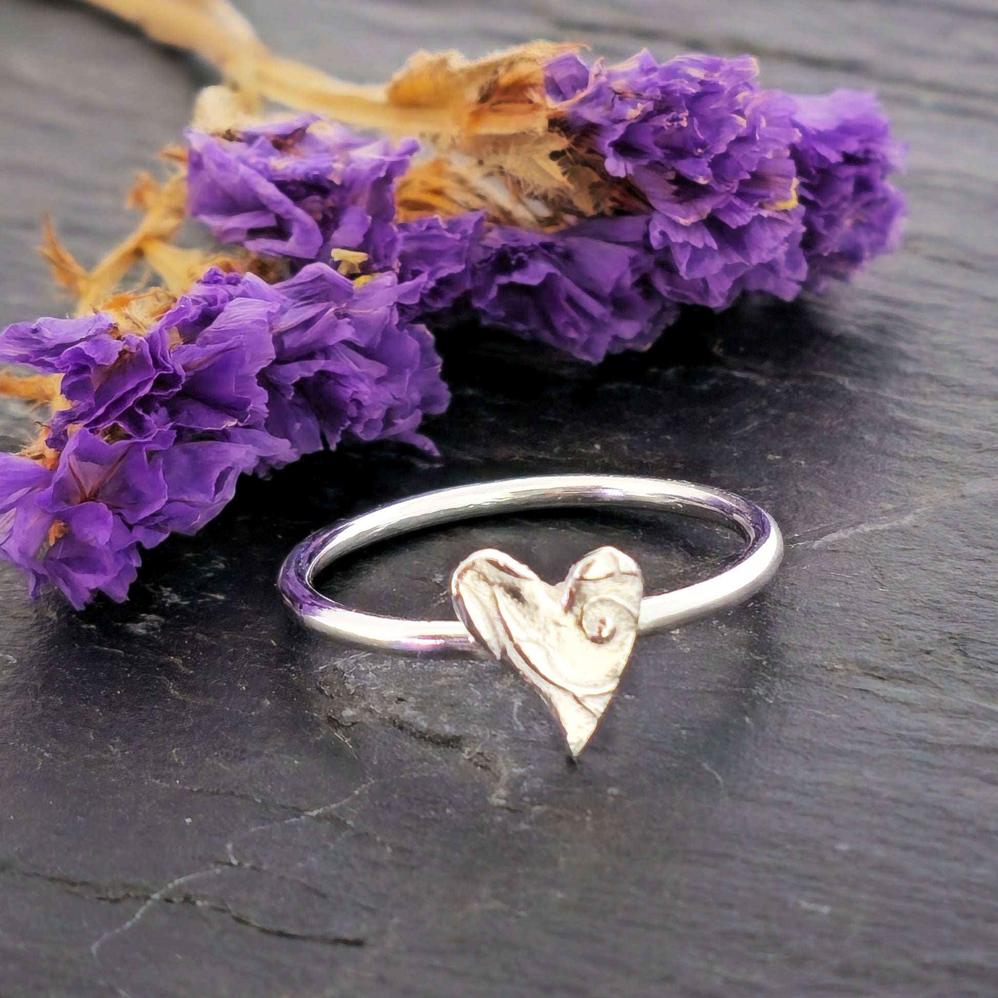 A handmade polished silver stacking ring with a textured heart charm resting on a dark slate surface next to dried purple flowers. Mijoux Jewellery.
