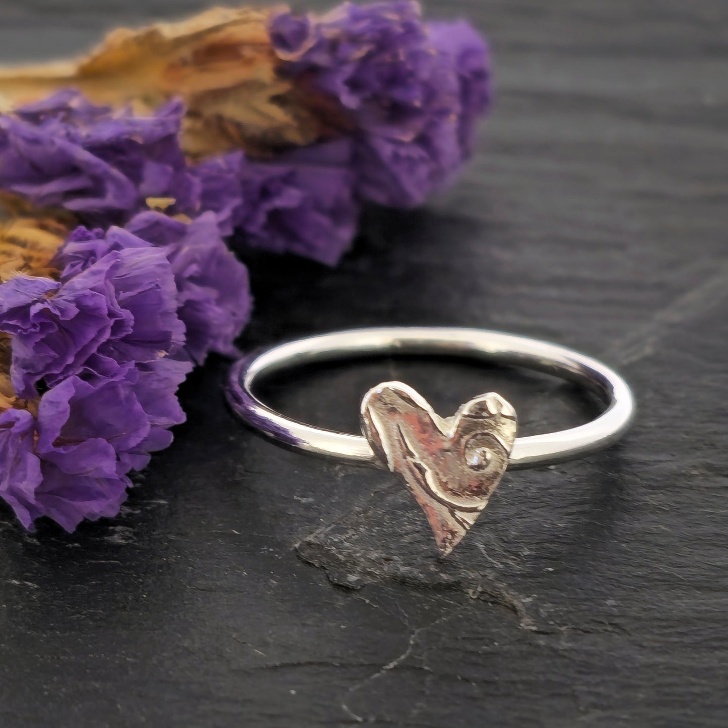 A handmade polished silver stacking ring with a textured heart charm resting on a dark slate surface next to dried purple flowers. Mijoux Jewellery.