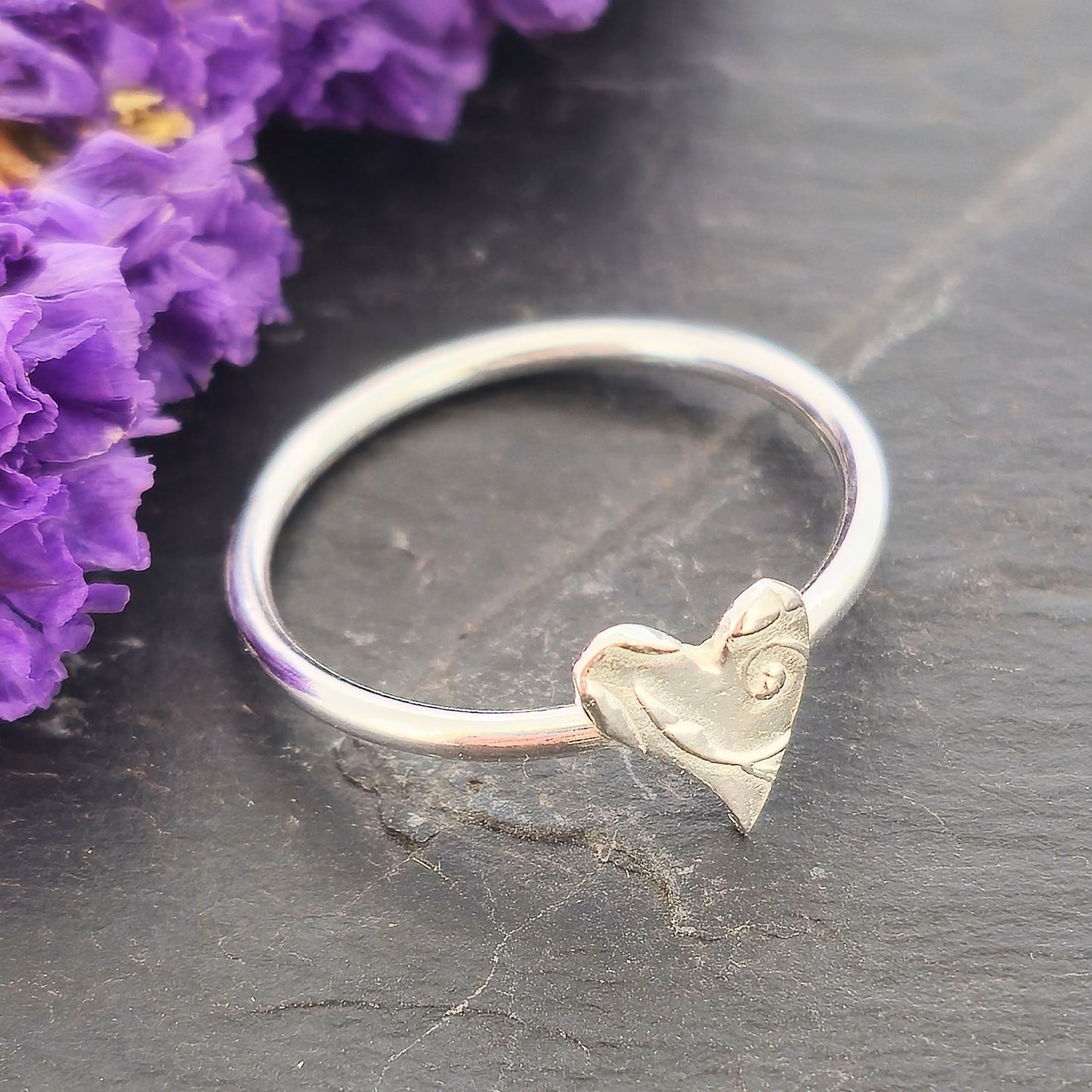 A handmade polished silver stacking ring with a textured heart charm resting on a dark slate surface next to dried purple flowers. Mijoux Jewellery.