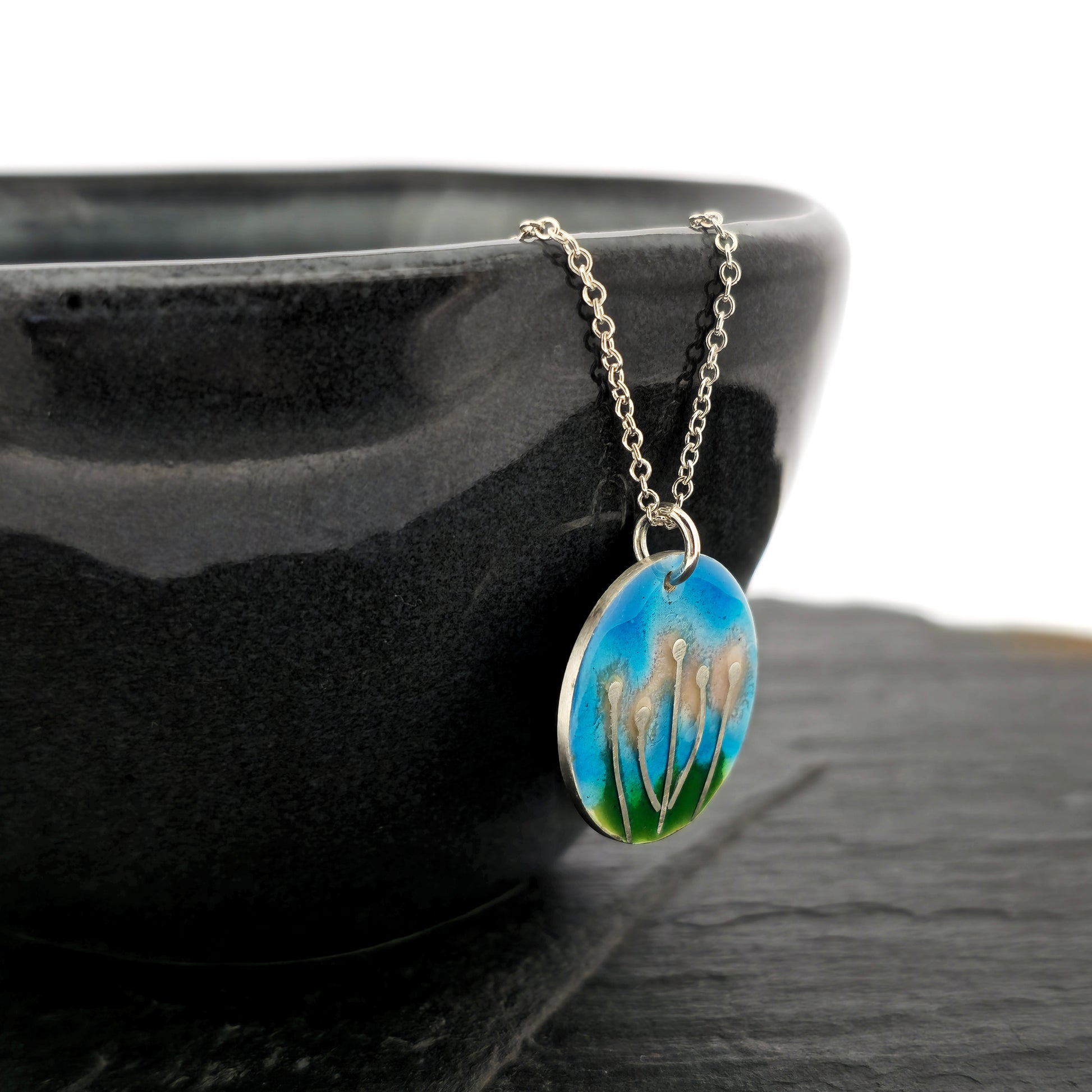 A round silver & enamel pendant with silver & pale pink flowers and a blue & green enamel background. On a silver chain. Side view. Shown on a black bowl.