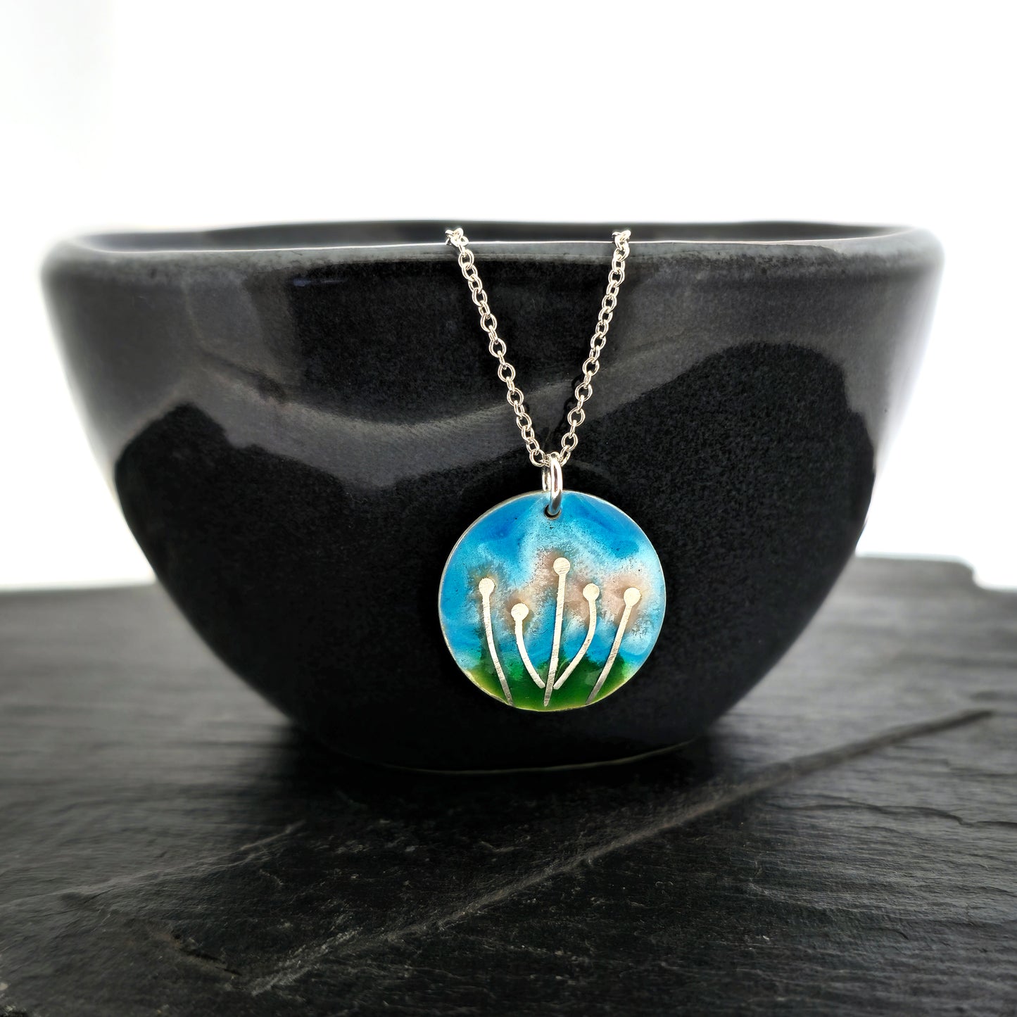 A round silver & enamel pendant with silver & pale pink flowers and a blue & green enamel background. On a silver chain. Shown on a black bowl.