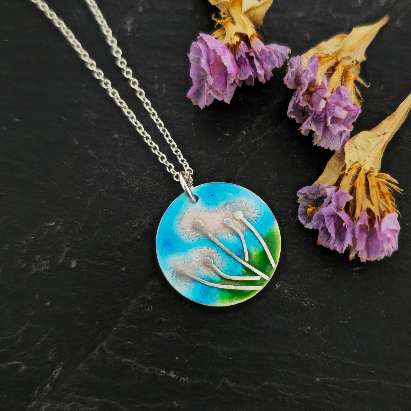 A round silver & enamel pendant with silver & pale pink flowers and a blue & green enamel background. On a silver chain. Shown on slate with flowers.