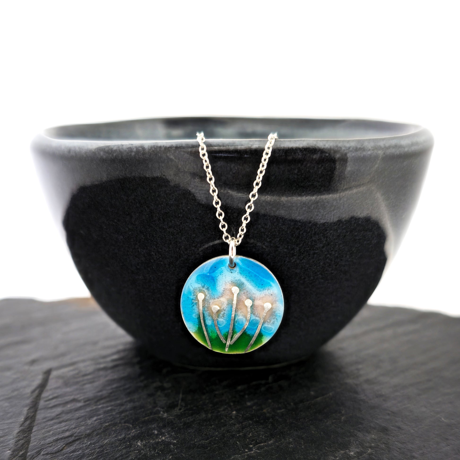 A round silver & enamel pendant with silver & pale pink flowers and a blue & green enamel background. On a silver chain. Shown on a black bowl.