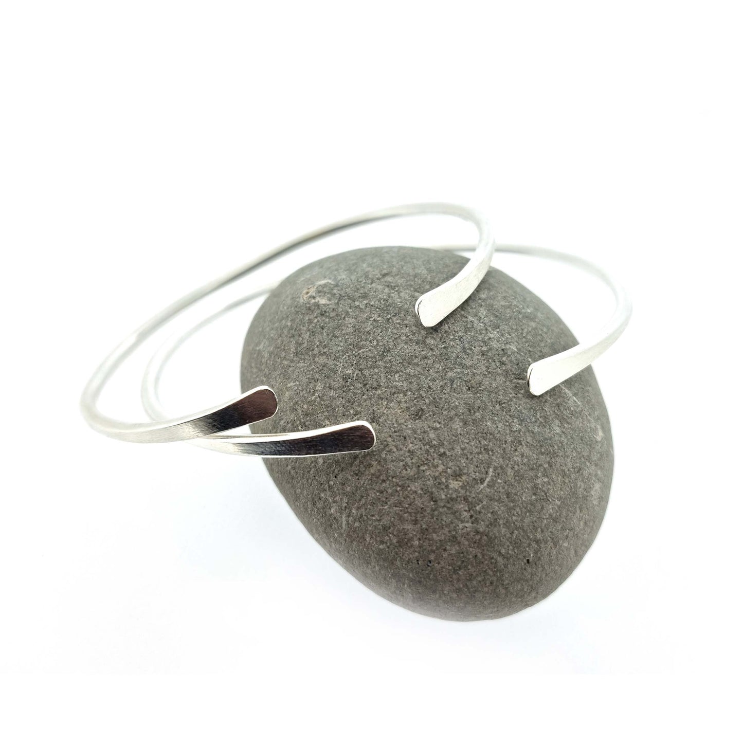 2 silver open torque bangles with flattened ends. Pictured on a pebble.