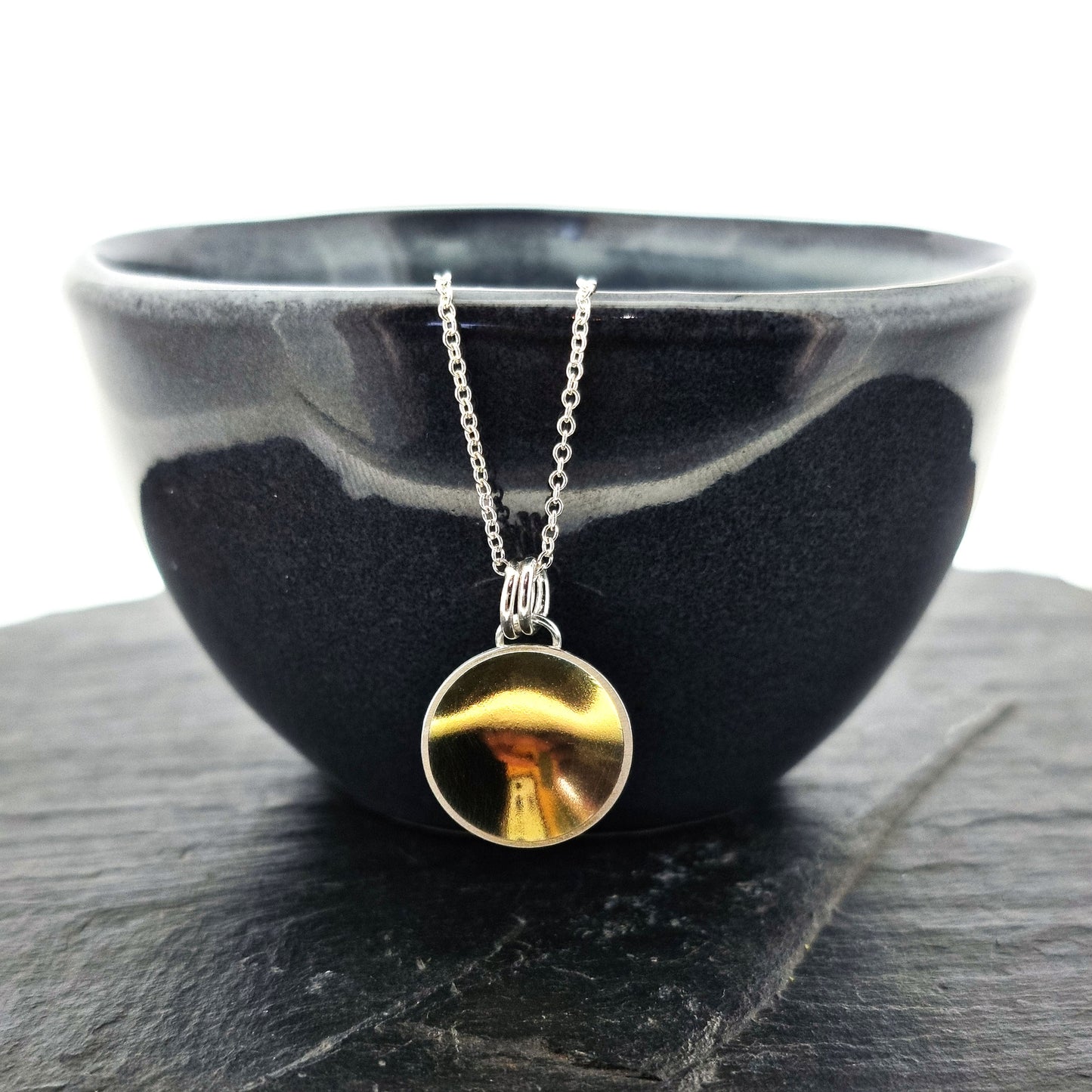 A silver round domed pendant with a gold center and double bail suspended from a silver chain. Pictured on a bowl.