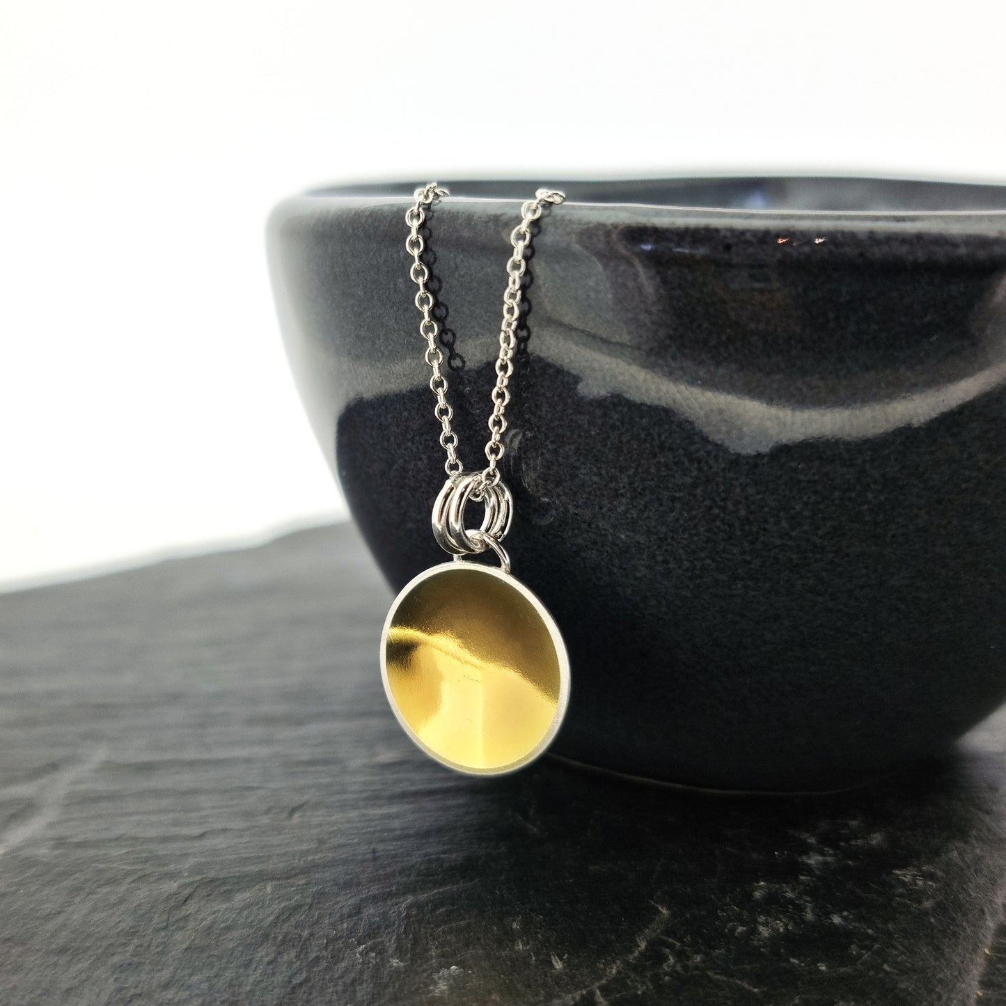 A silver round domed pendant with a gold center and double bail suspended from a silver chain. Pictured on a bowl.