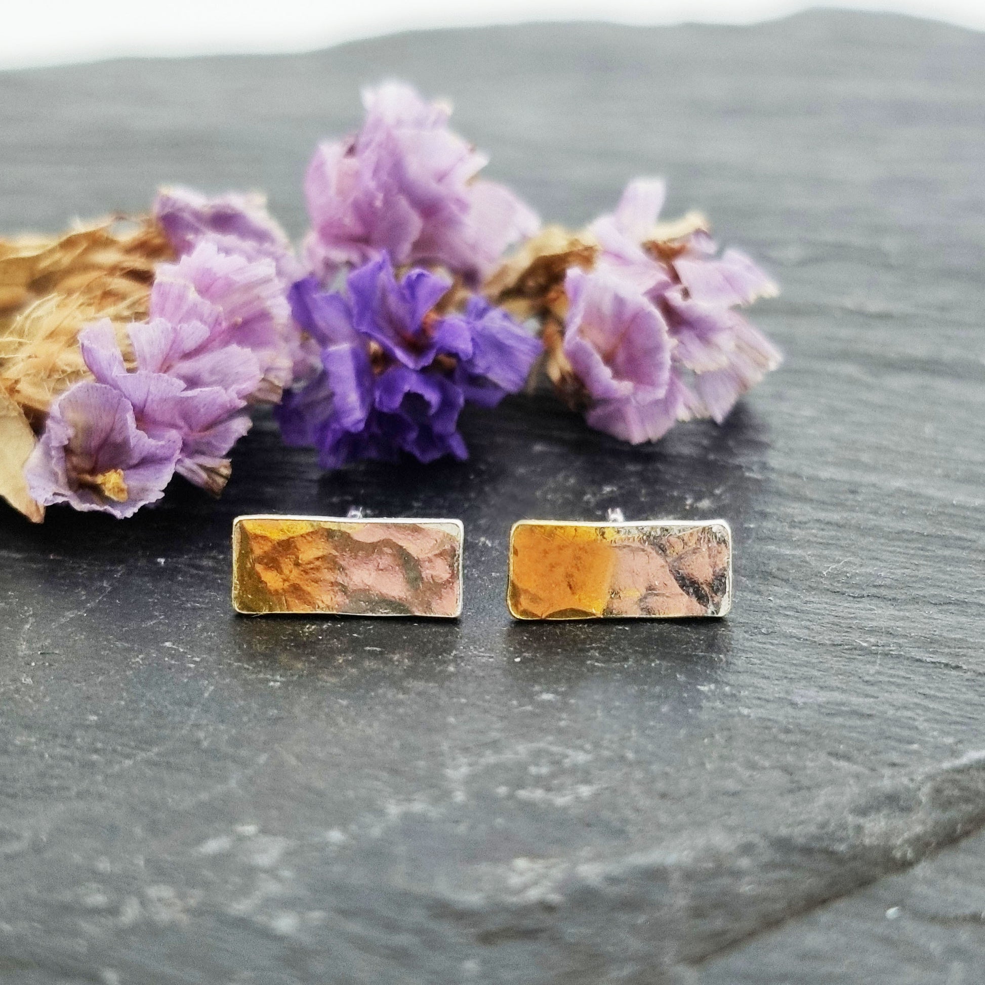 Silver rectangular stud earrings with a hammered finish and one half gold plated. Pictured with flowers.