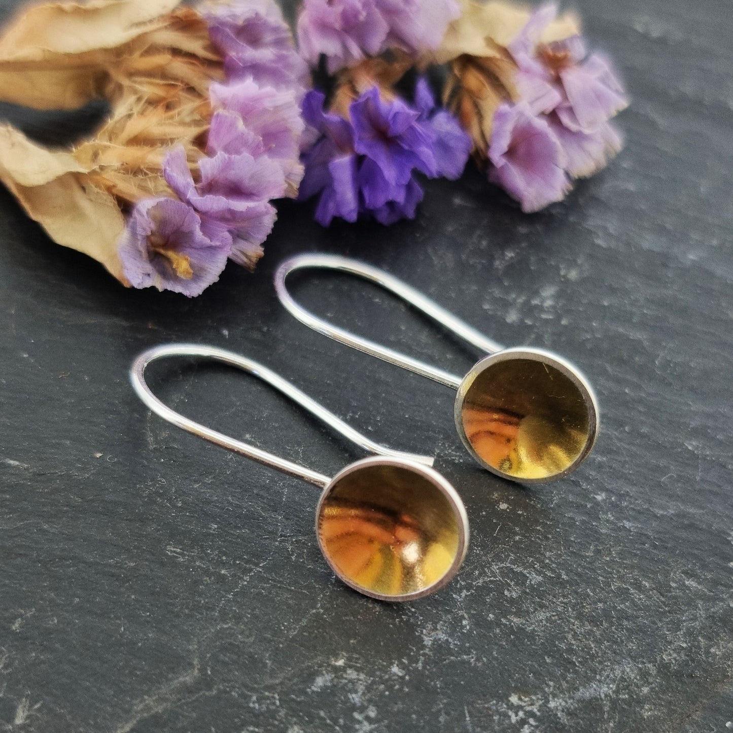 Silver drop earrings with a domed round section with a gold center suspended from a silver ear wire. Pictured with flowers.