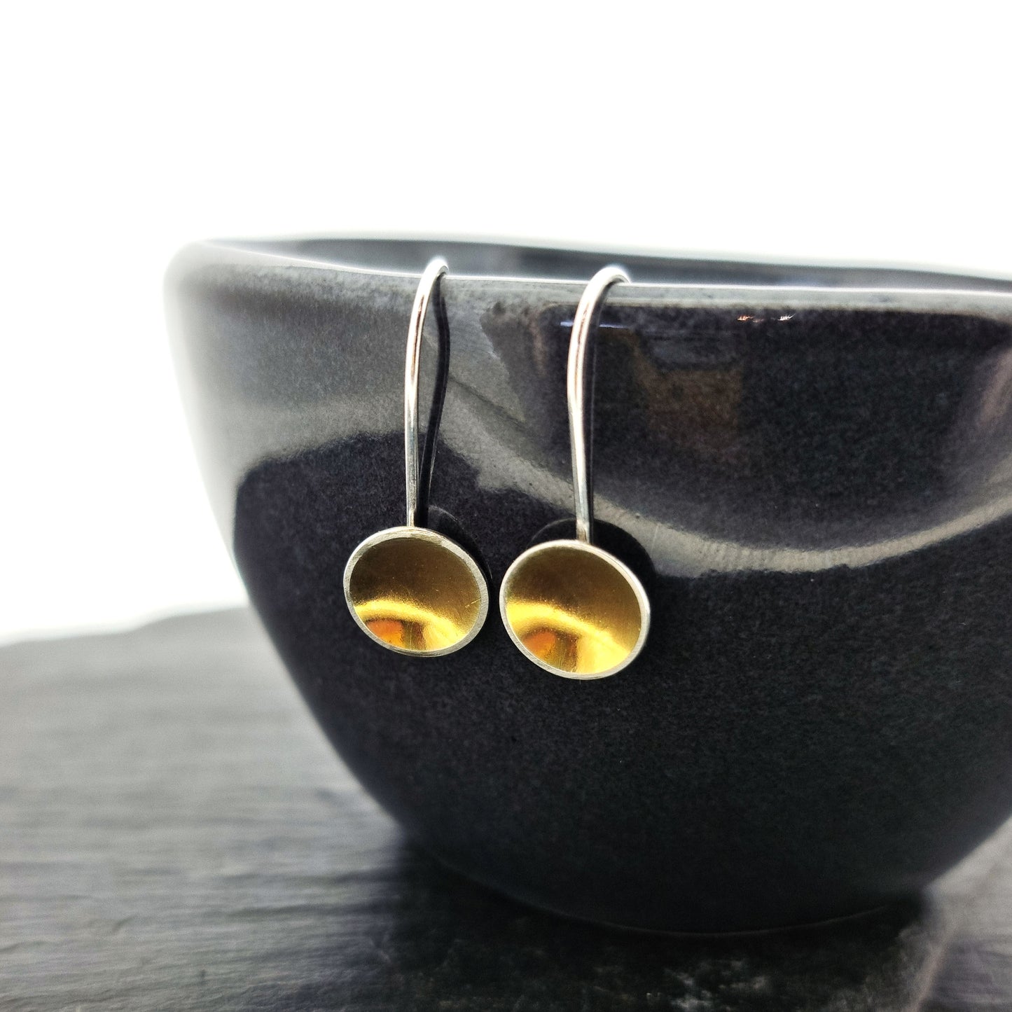 Silver drop earrings with a domed round section with a gold center suspended from a silver ear wire. Pictured on a bowl.