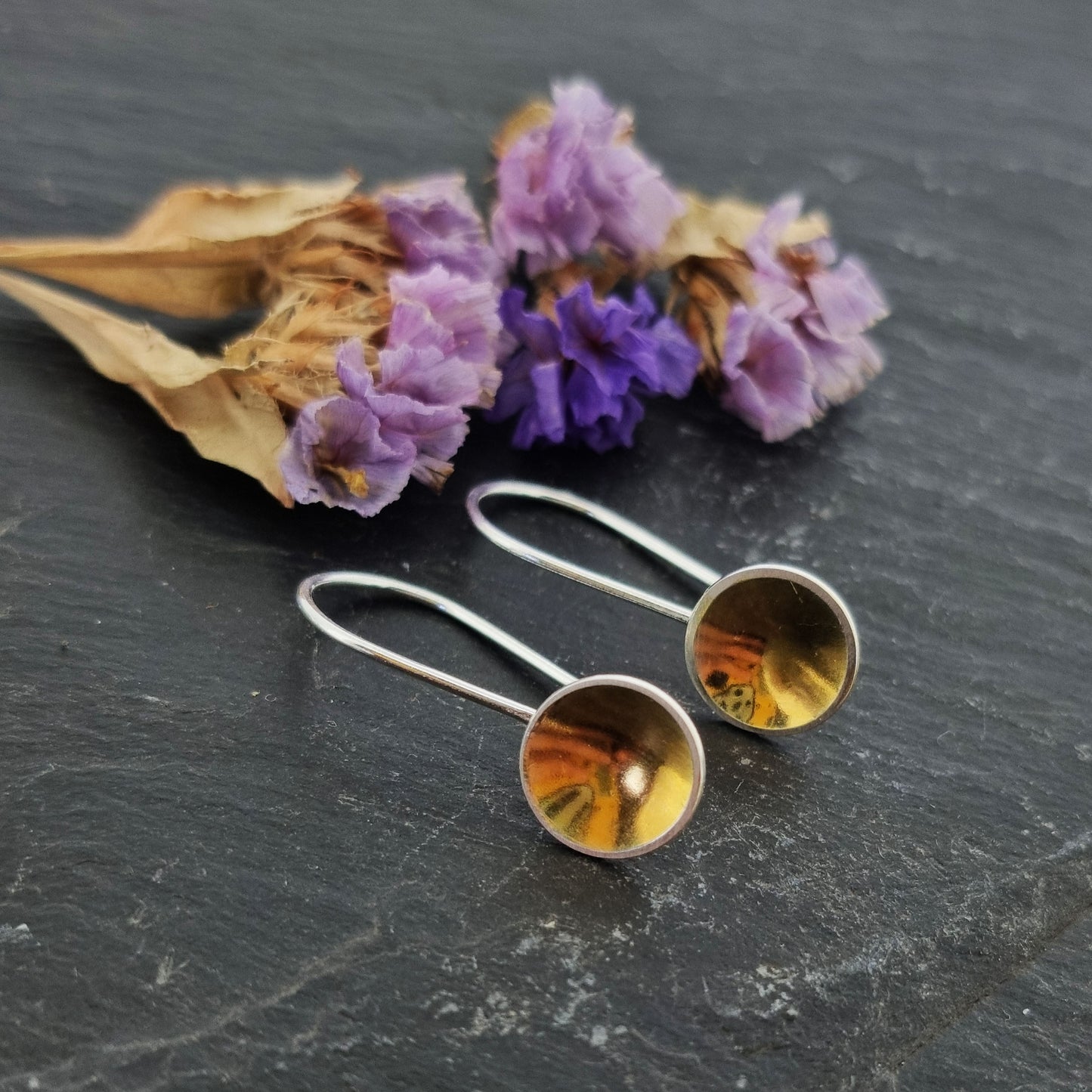 Silver drop earrings with a domed round section with a gold center suspended from a silver ear wire. Pictured with flowers.