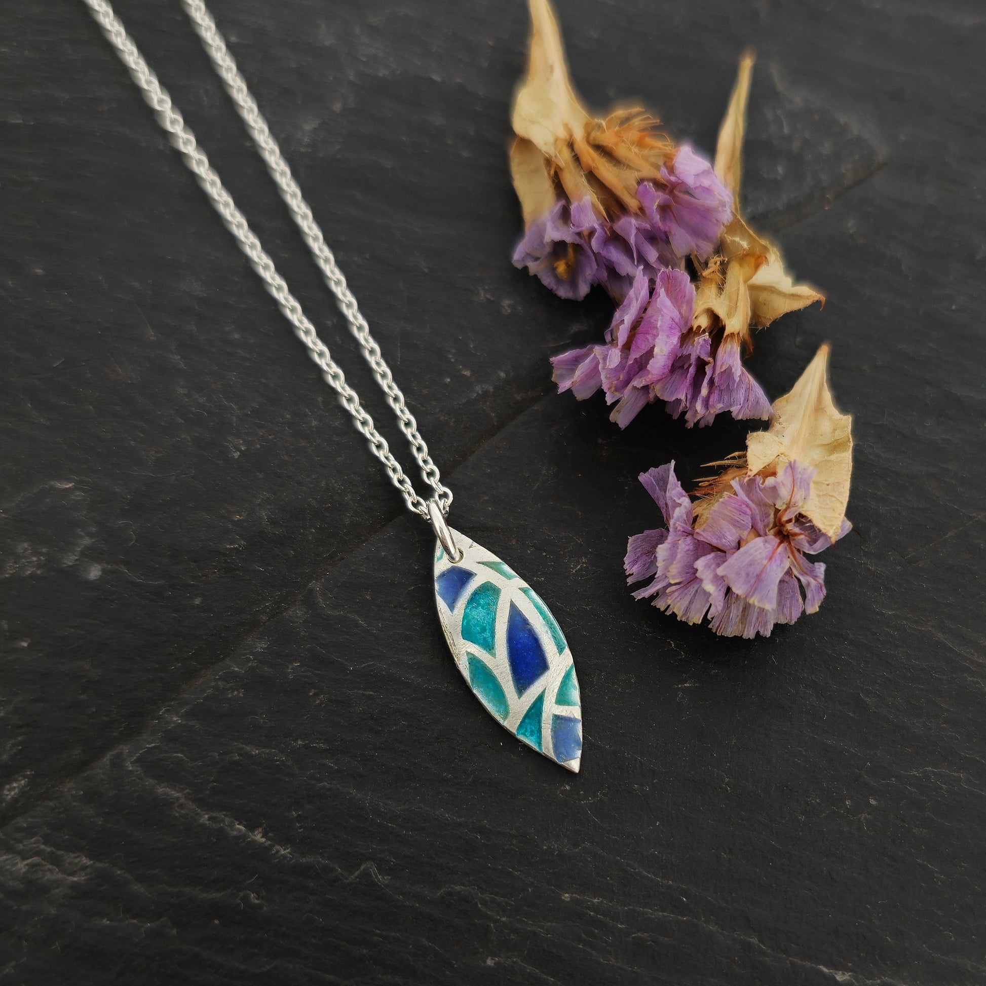 A marquise shaped silver pendant with geometric cells of blue, green and turquoise enamel. On a silver chain. Small. Pictured on slate with flowers.