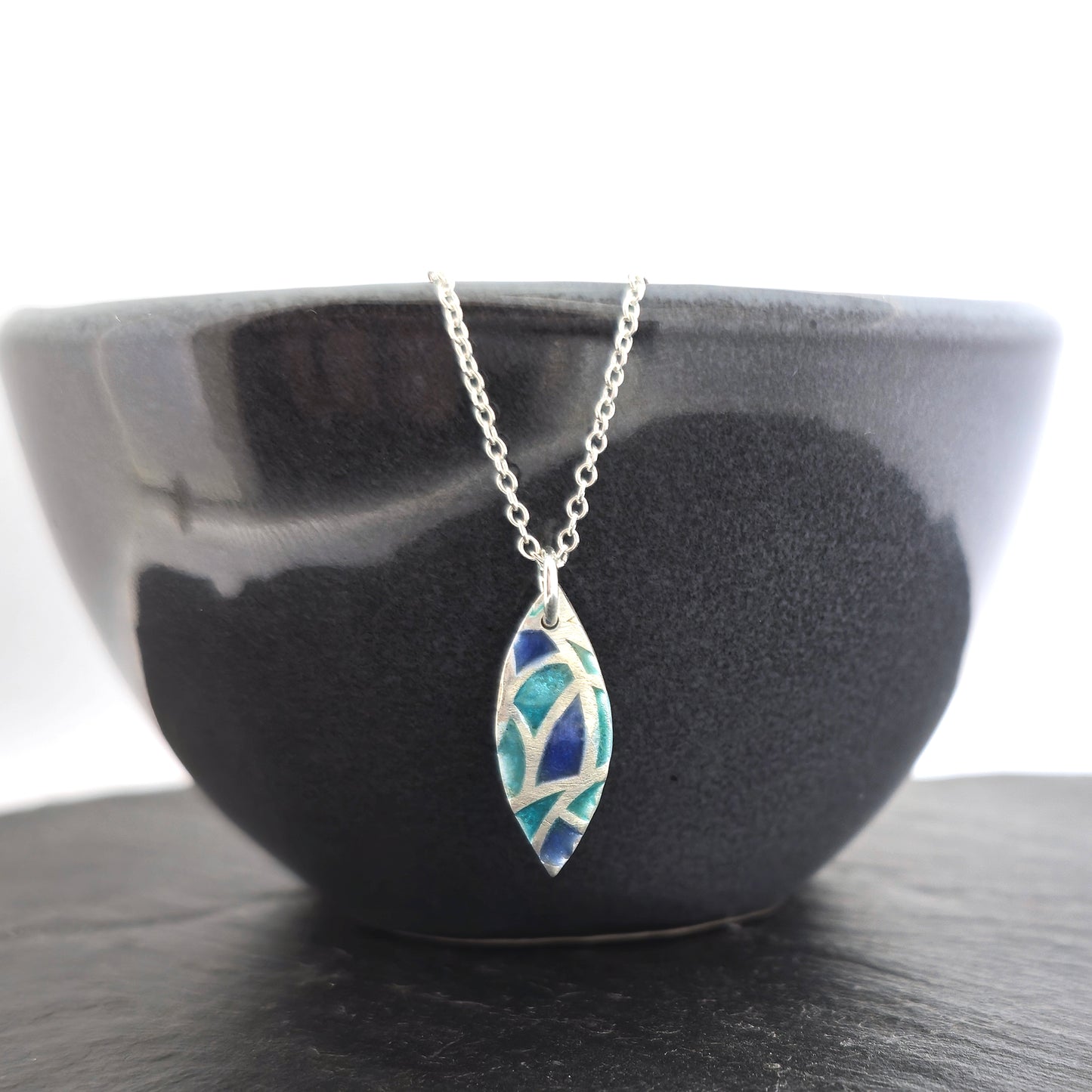 A marquise shaped silver pendant with geometric cells of blue, green and turquoise enamel. On a silver chain. Small. Pictured on a black bowl.