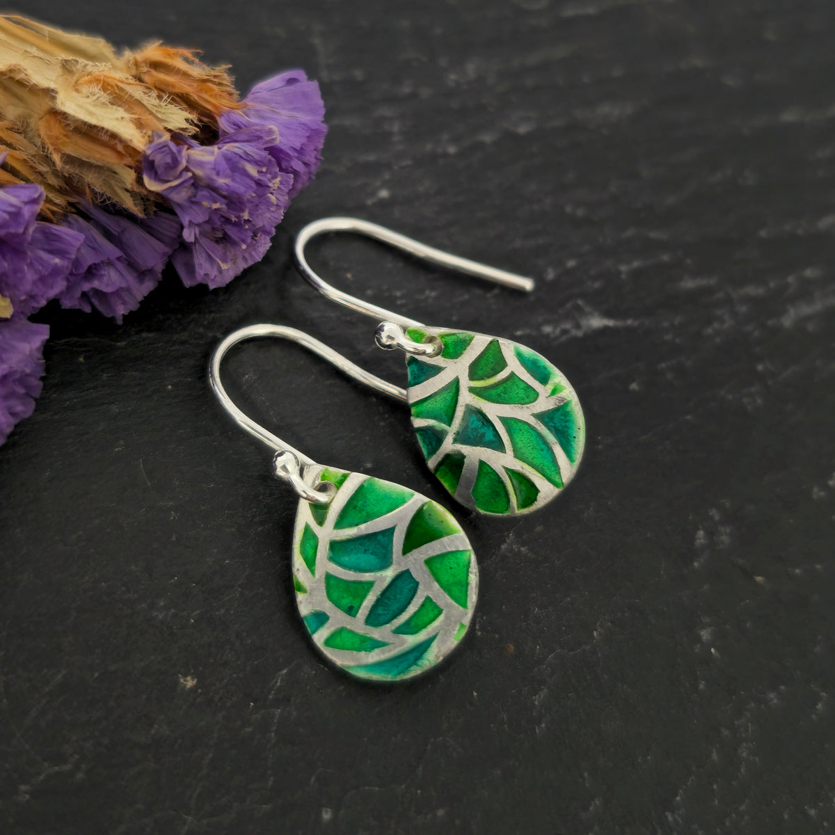 A pair of handmade silver teardrop dangle earrings with green enamel mosaic leaf designs, displayed on a dark textured surface next to dried purple flowers. Mijoux Jewellery.