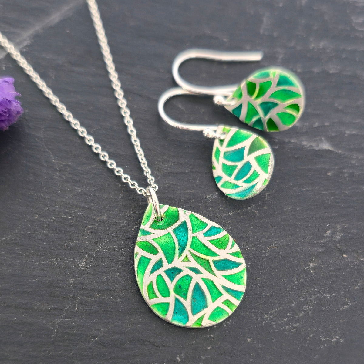 A handmade silver necklace and matching drop earrings with teardrop-shaped pendants featuring a vibrant green enamel mosaic leaf pattern, displayed on a dark slate surface. Mijoux Jewellery.