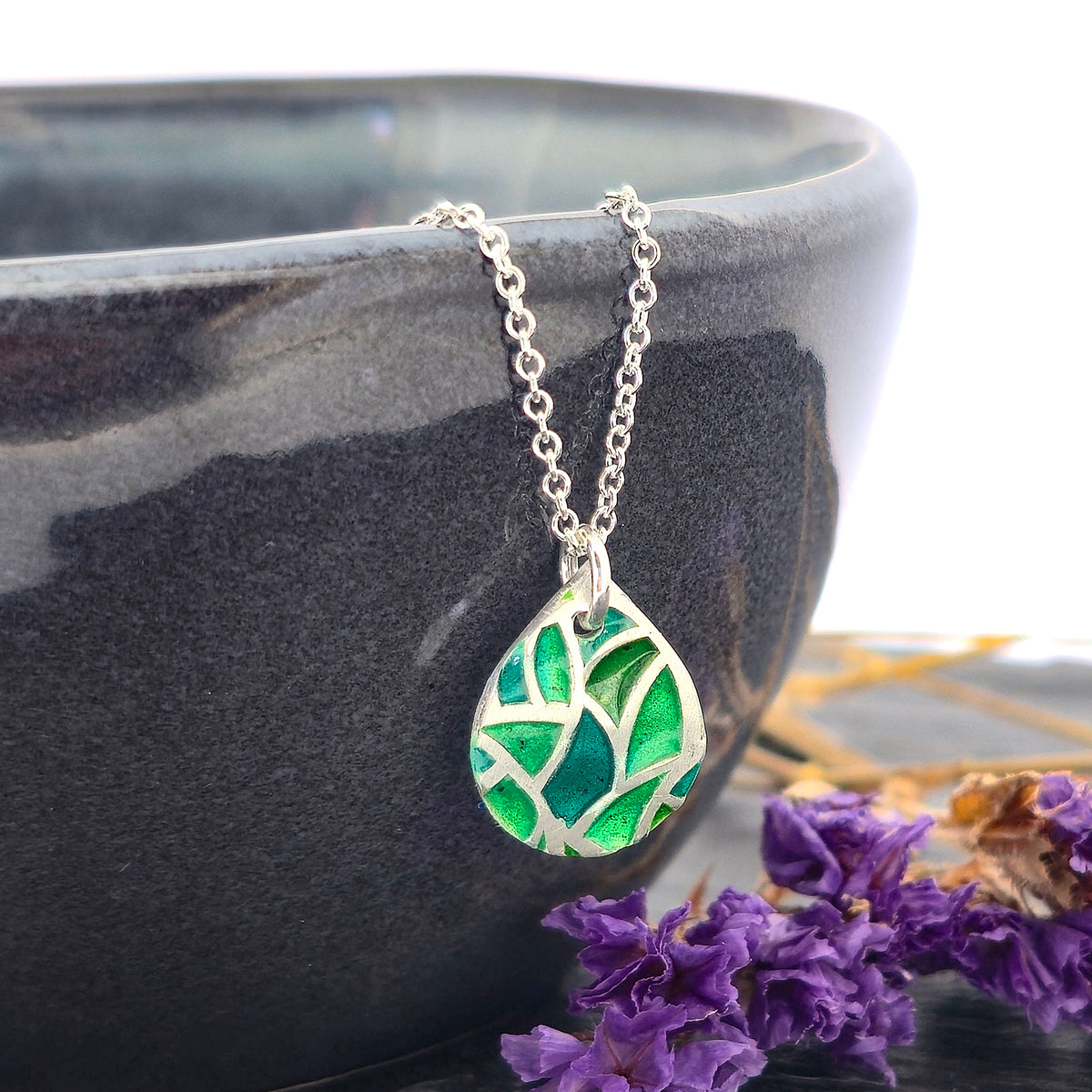 A handmade teardrop-shaped pendant necklace featuring a green enamel mosaic pattern with a silver chain, shown draped on a black bowl with dried purple flowers. Mijoux Jewellery.