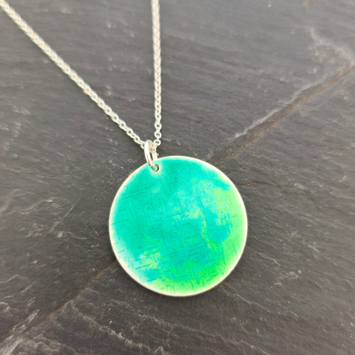 A silver chain necklace with a handmade round silver disc pendant featuring a vibrant green and turquoise textured pattern, displayed on a dark slate surface. Mijoux Jewellery.