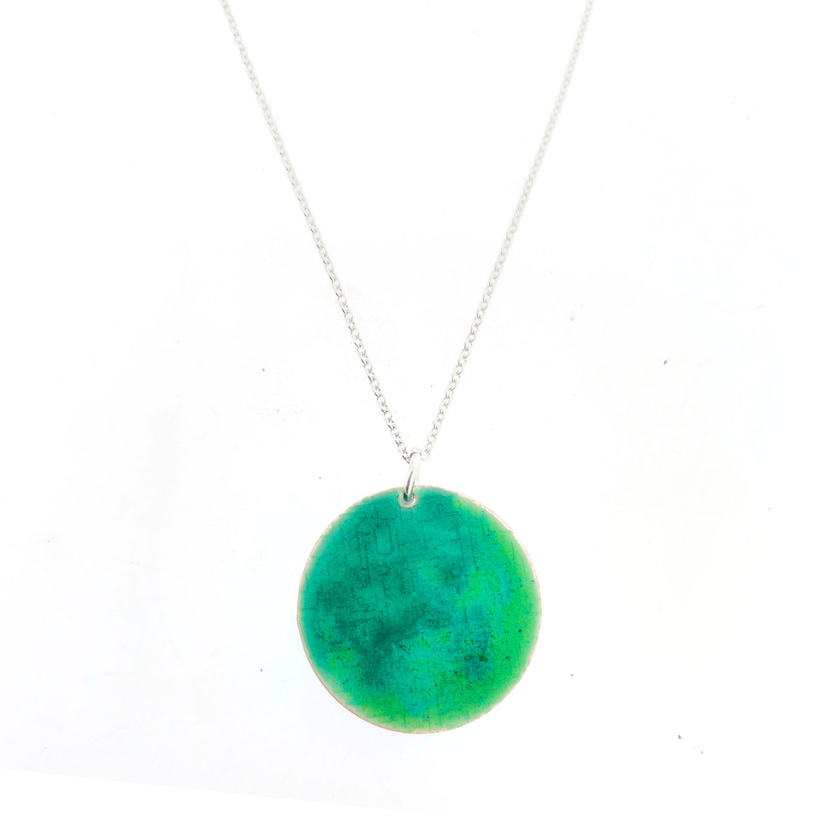 A handmade disc pendant with a textured pattern and a mix of green and turquoise enamel, suspended on a silver chain. On a white background. Mijoux Jewellery.