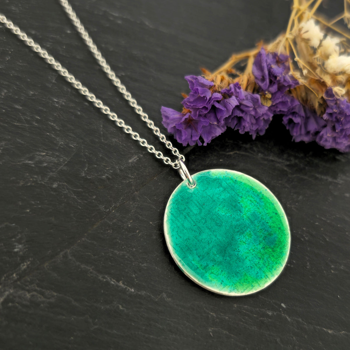 A silver chain necklace with a handmade round silver disc pendant featuring a vibrant green and turquoise enamelled textured pattern, displayed on a dark slate surface. Mijoux Jewellery.