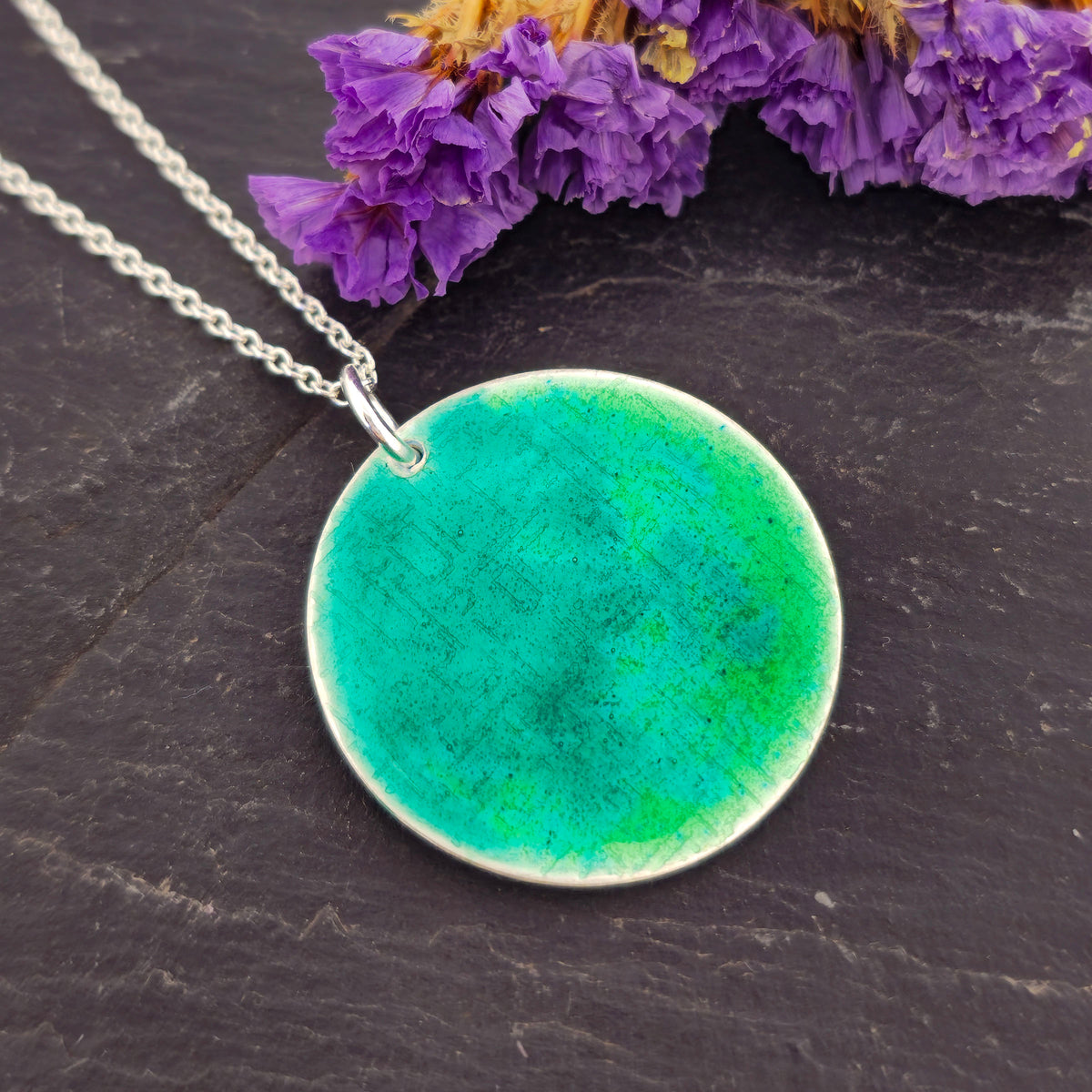 A silver chain necklace with a handmade round silver disc pendant featuring a vibrant green and turquoise enamelled textured pattern, displayed on a dark slate surface. Mijoux Jewellery.
