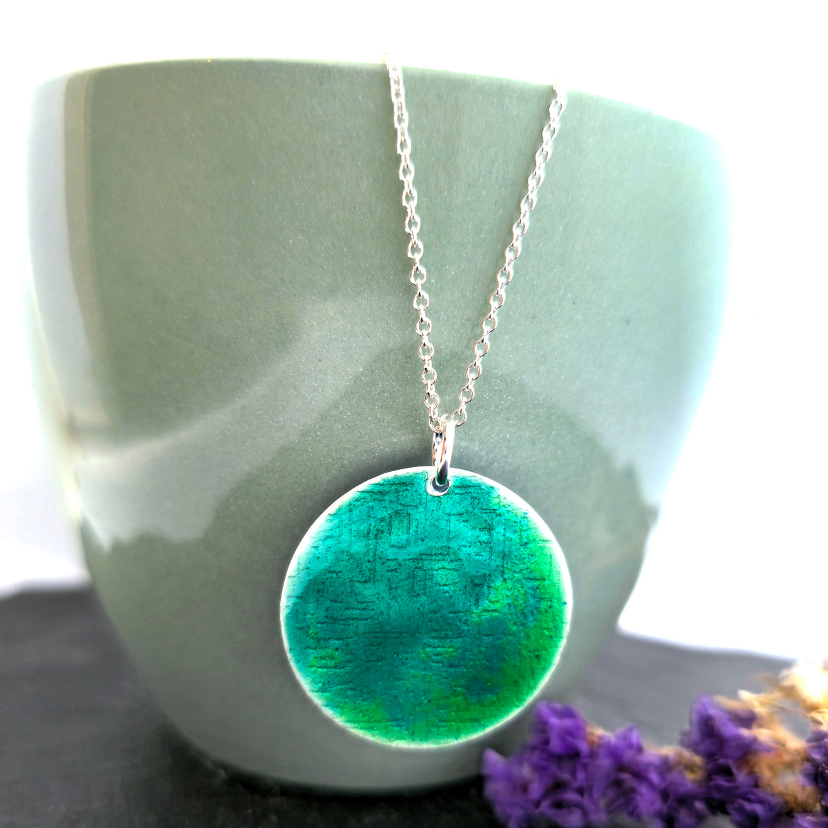 A silver chain necklace with a handmade silver round pendant featuring a green and teal marbled enamel pattern, displayed draped over a light green ceramic mug with dried purple flowers on a dark surface. Mijoux Jewellery.