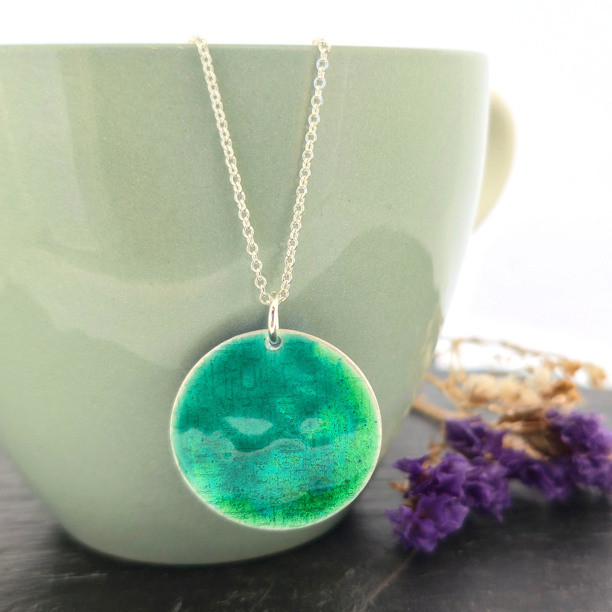 A silver chain necklace with a handmade silver round pendant featuring a green and teal marbled enamel pattern, displayed draped over a light green ceramic mug with dried purple flowers on a dark surface. Mijoux Jewellery.
