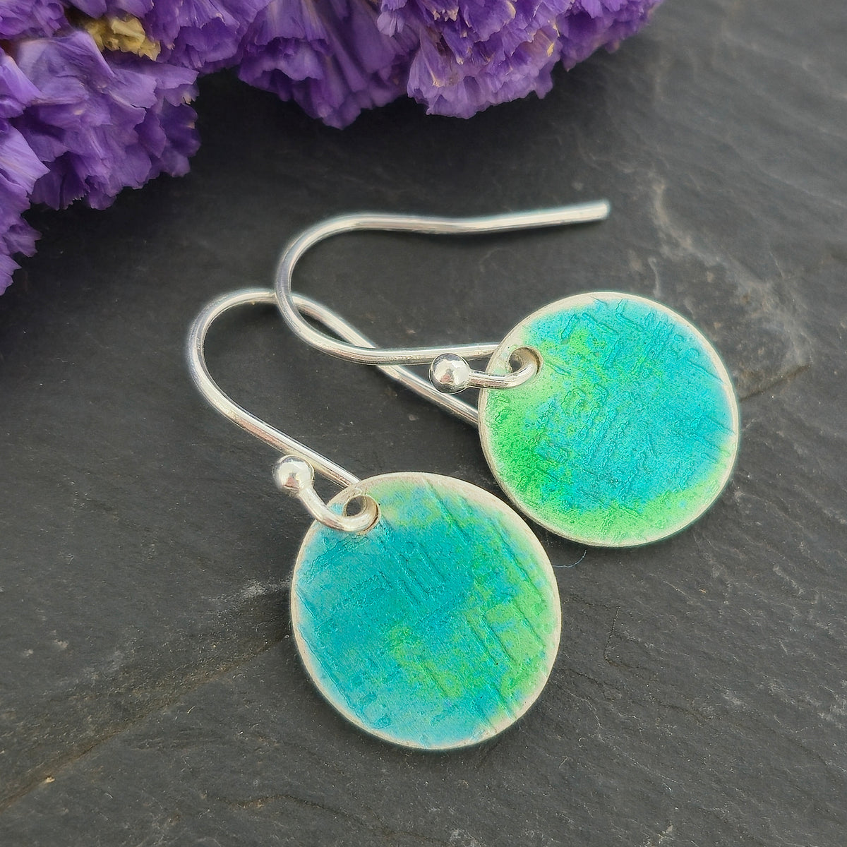A pair of handmade silver fishhook earrings with round, textured disc charms in a vibrant turquoise and green gradient enamel, displayed on a dark slate surface with purple flowers in the background. Mijoux Jewellery.