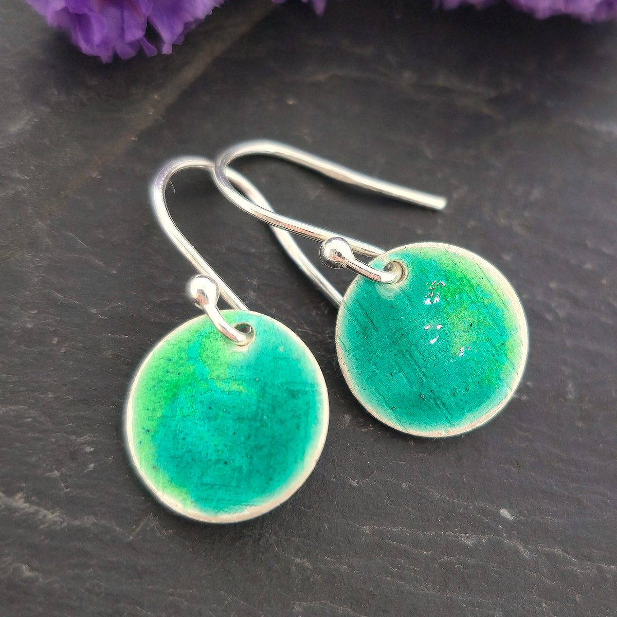 A pair of handmade silver fishhook earrings with round, textured disc charms in a vibrant turquoise and green gradient enamel, displayed on a dark slate surface with purple flowers in the background. Mijoux Jewellery.
