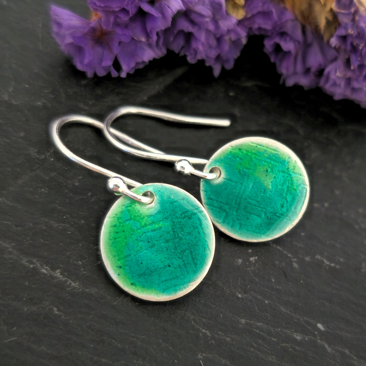 A pair of handmade silver fishhook earrings with round, textured disc charms in a vibrant turquoise and green gradient enamel, displayed on a dark slate surface with purple flowers in the background. Mijoux Jewellery.
