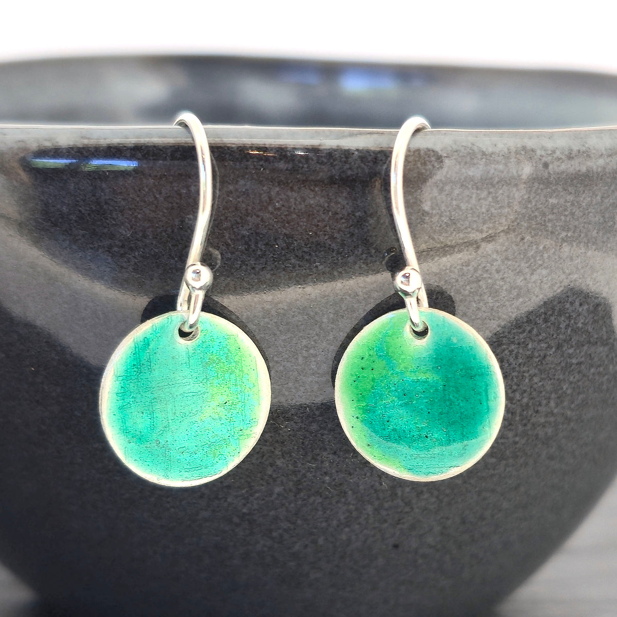 A pair of handmade silver fishhook earrings with round, textured disc charms in a vibrant turquoise and green gradient enamel, displayed on a dark bowl. Mijoux Jewellery.