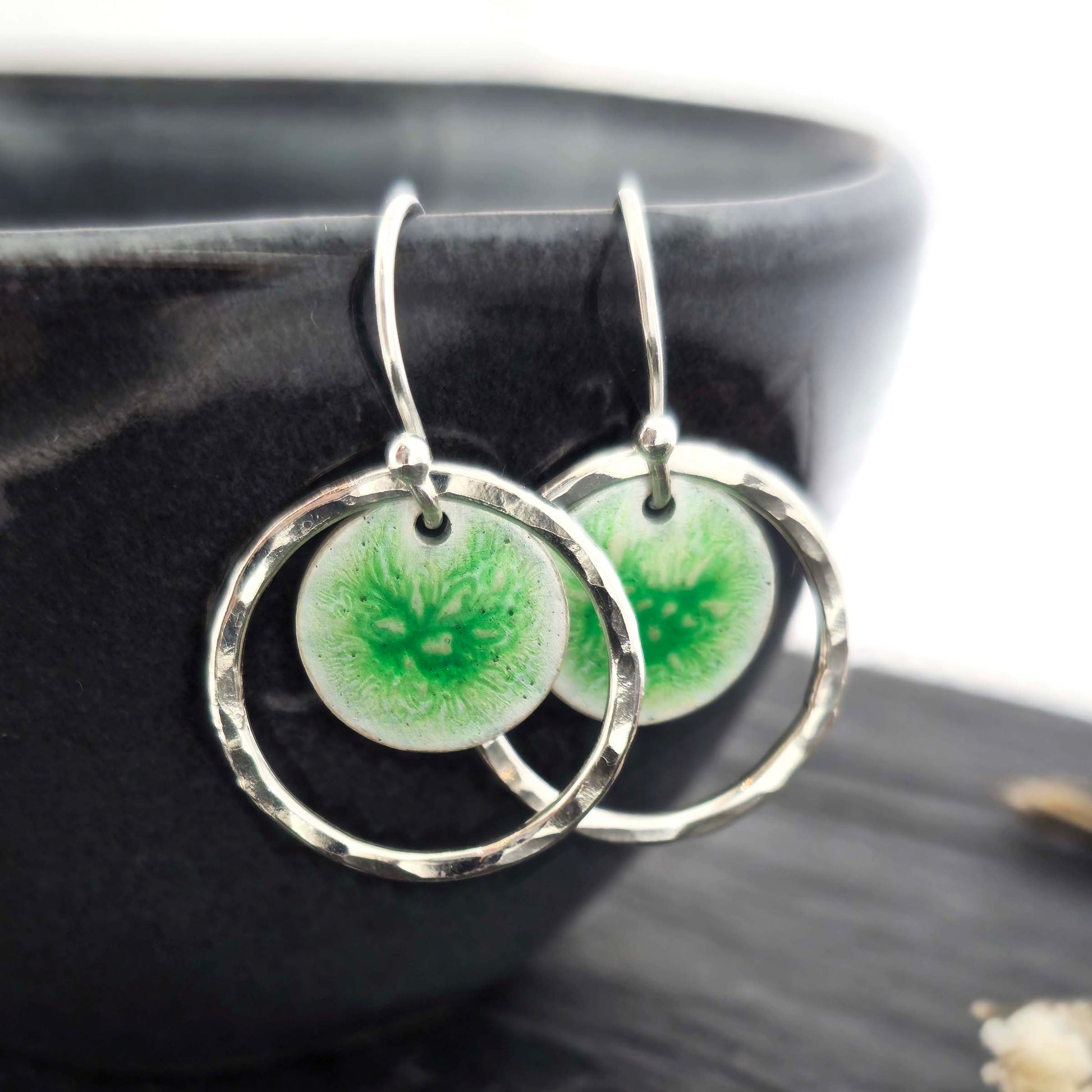 Silver drop earrings with a central green patterned enamel disc and a surrounding hammered silver circle. Shown on a black bowl.