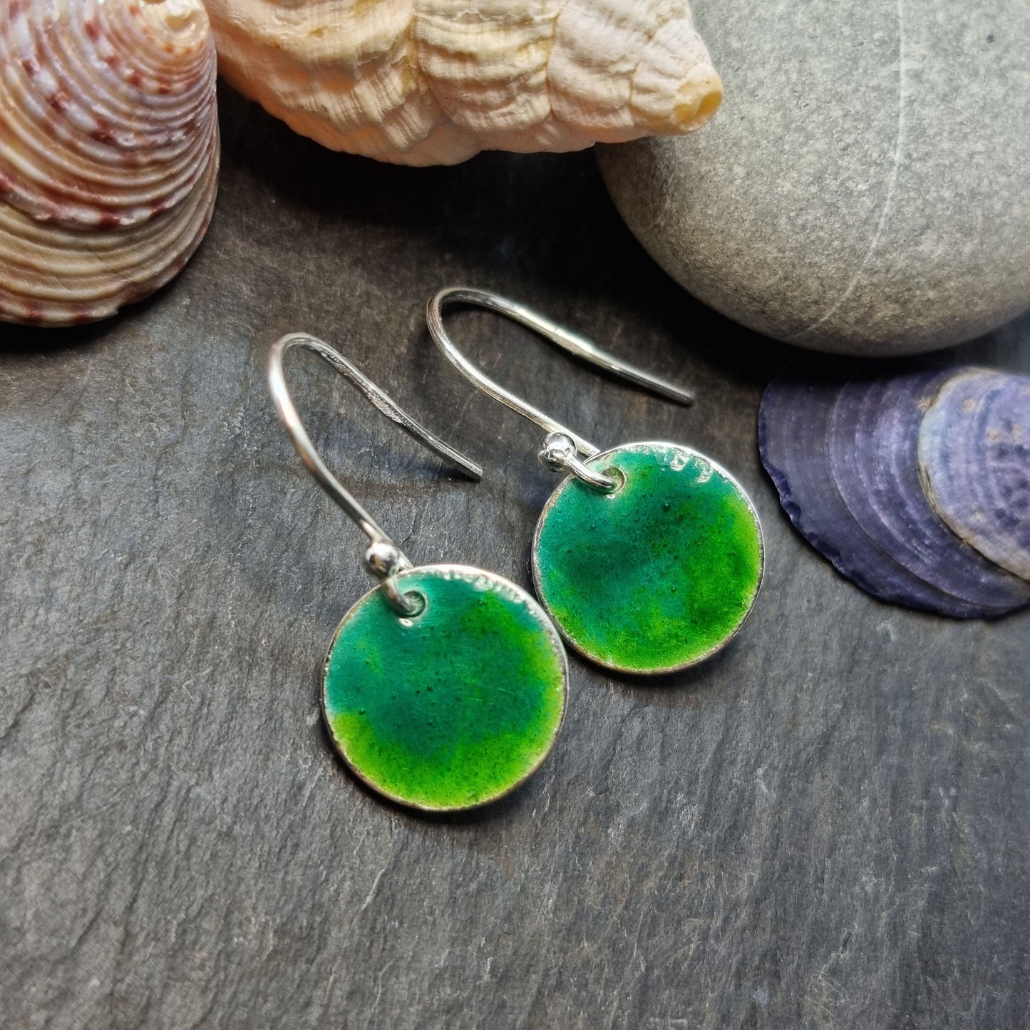 Round silver drop earrings with a mix of green enamels. Pictured with pebbles and shells.