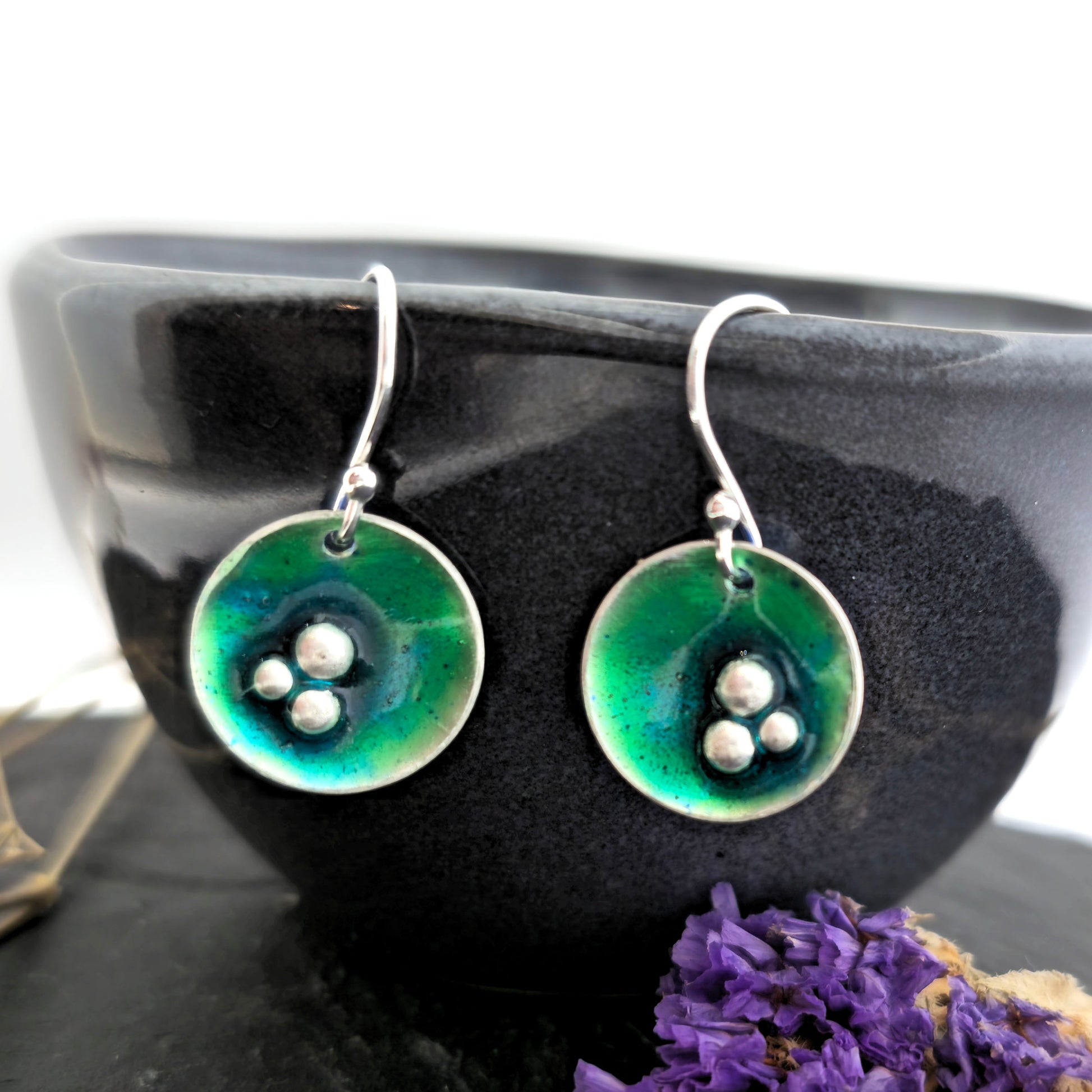 Handmade silver round drop earrings with green & turquoise enamel and 3 silver balls off centre. On silver ear hooks. Shown on a black bowl with flowers. Mijoux Jewellery.
