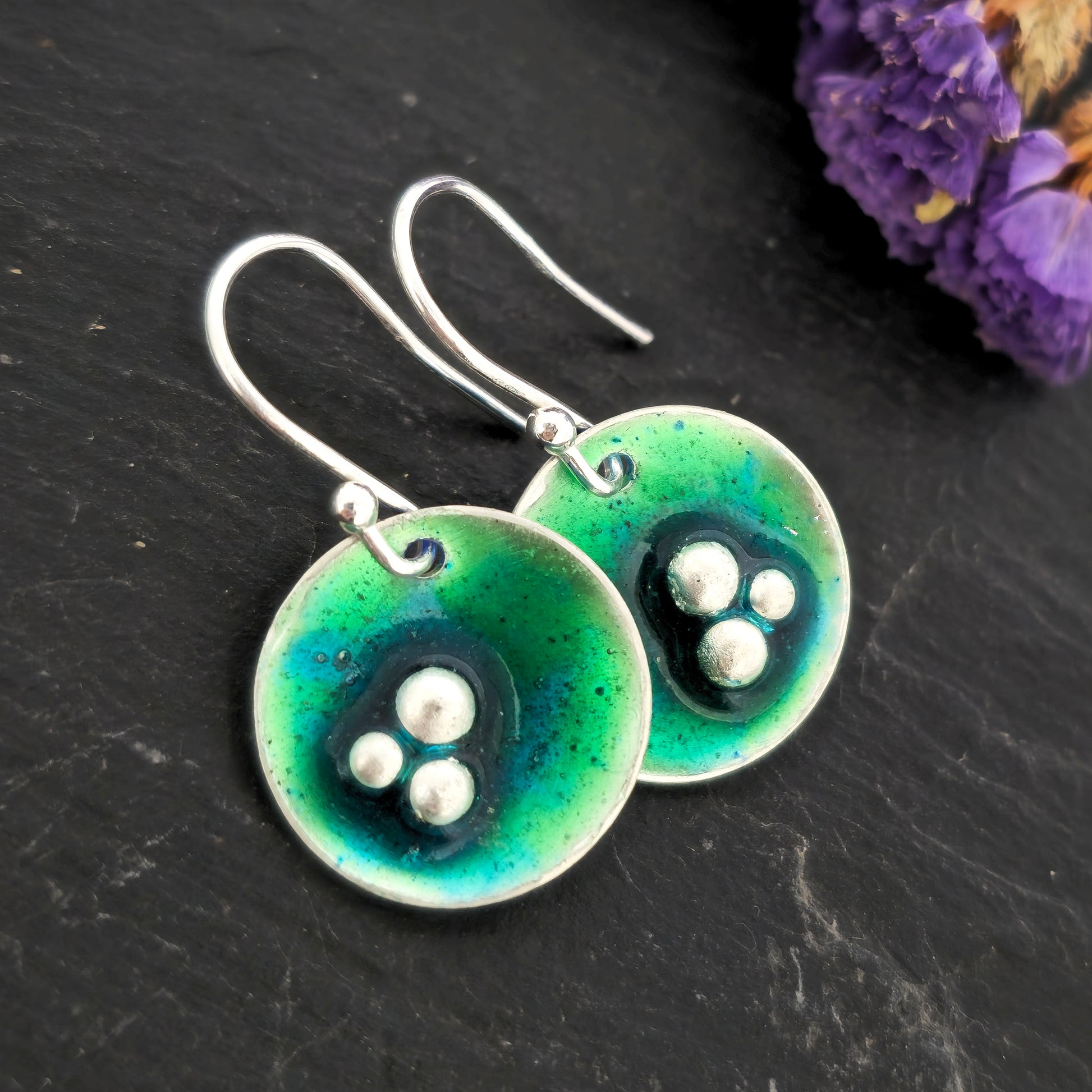 Handmade silver round drop earrings with green & turquoise enamel and 3 silver balls off centre. On silver ear hooks. Shown on a black background with flowers. Mijoux Jewellery.
