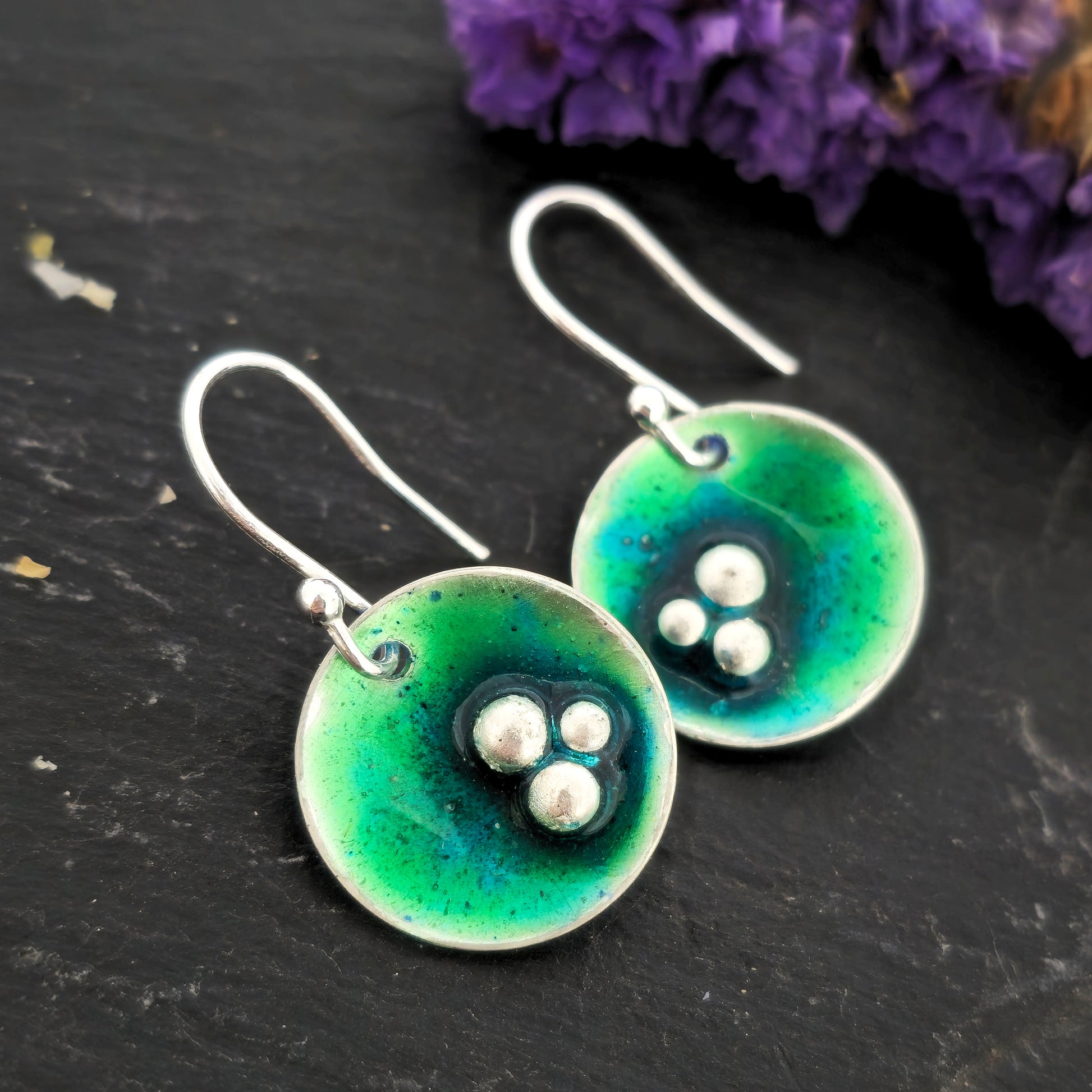 Handmade silver round drop earrings with green & turquoise enamel and 3 silver balls off centre. On silver ear hooks. Shown on a black background with flowers. Mijoux Jewellery.