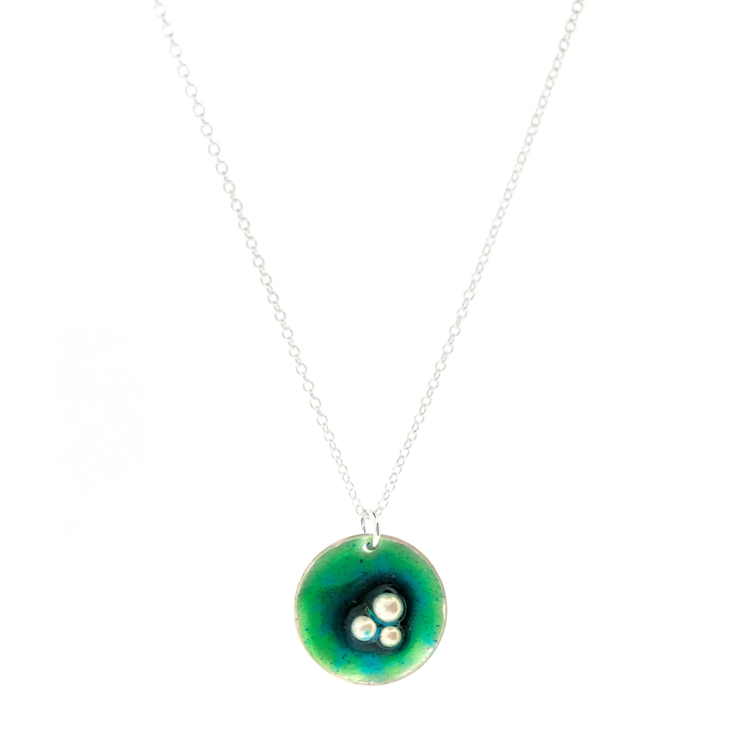 A handmade round silver pendant with a mix of green and turquoise enamel and three silver balls off-centre. Suspended on a silver chain. Shown on a white background. Mijoux Jewellery.
