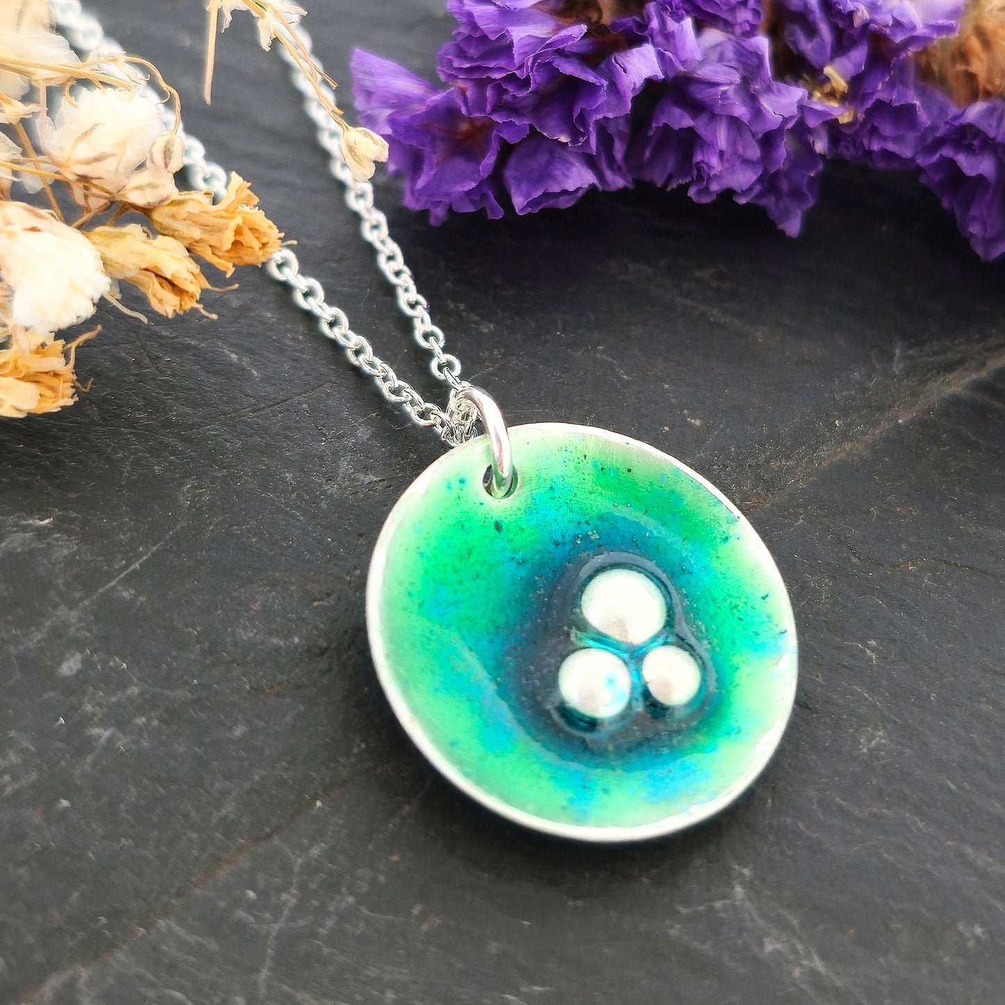 A handmade round silver pendant with a mix of green and turquoise enamel and three silver balls off-centre. Suspended on a silver chain. Shown on a black background with flowers. Mijoux Jewellery.