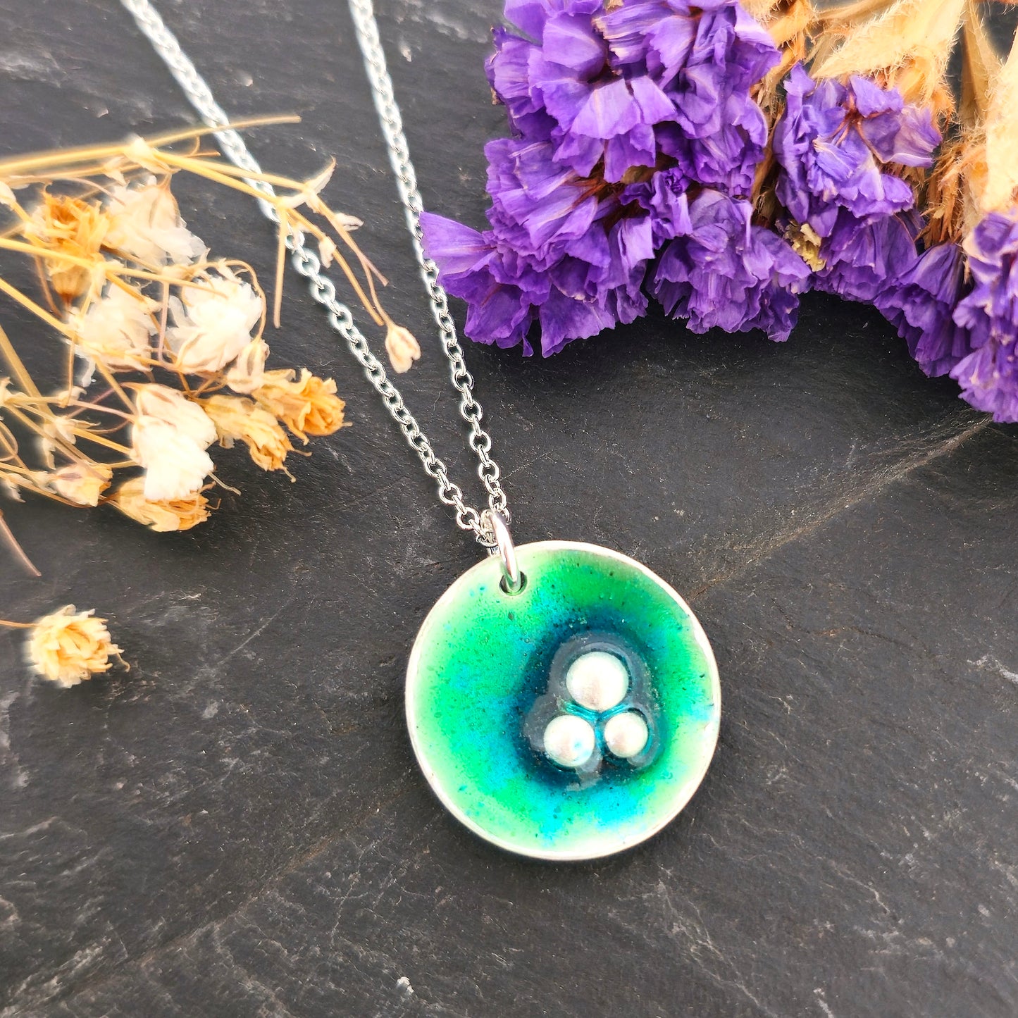 A handmade round silver pendant with a mix of green and turquoise enamel and three silver balls off-centre. Suspended on a silver chain. Shown on a black background with flowers. Mijoux Jewellery.