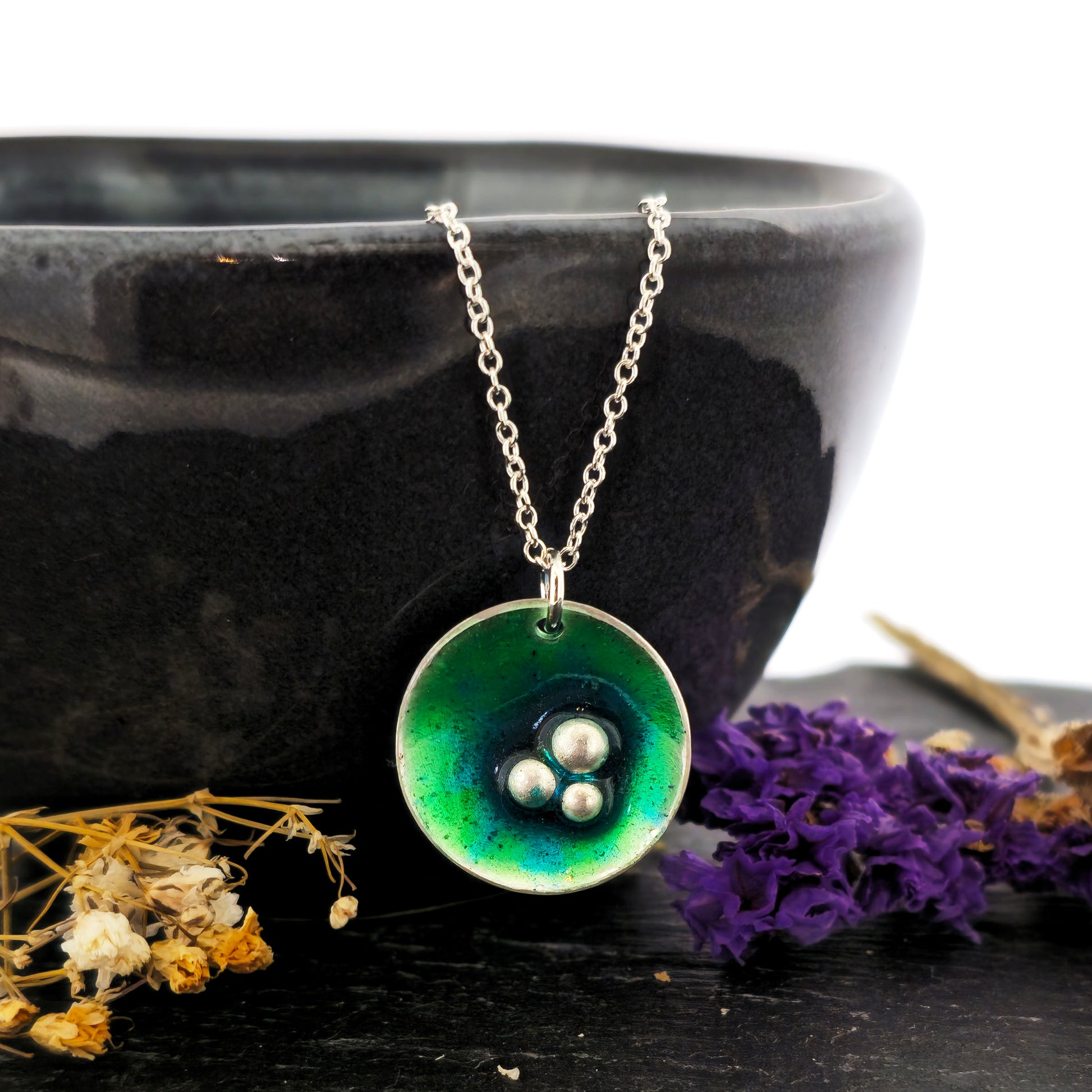 A handmade round silver pendant with a mix of green and turquoise enamel and three silver balls off-centre. Suspended on a silver chain. Shown on a black background with flowers. Mijoux Jewellery.