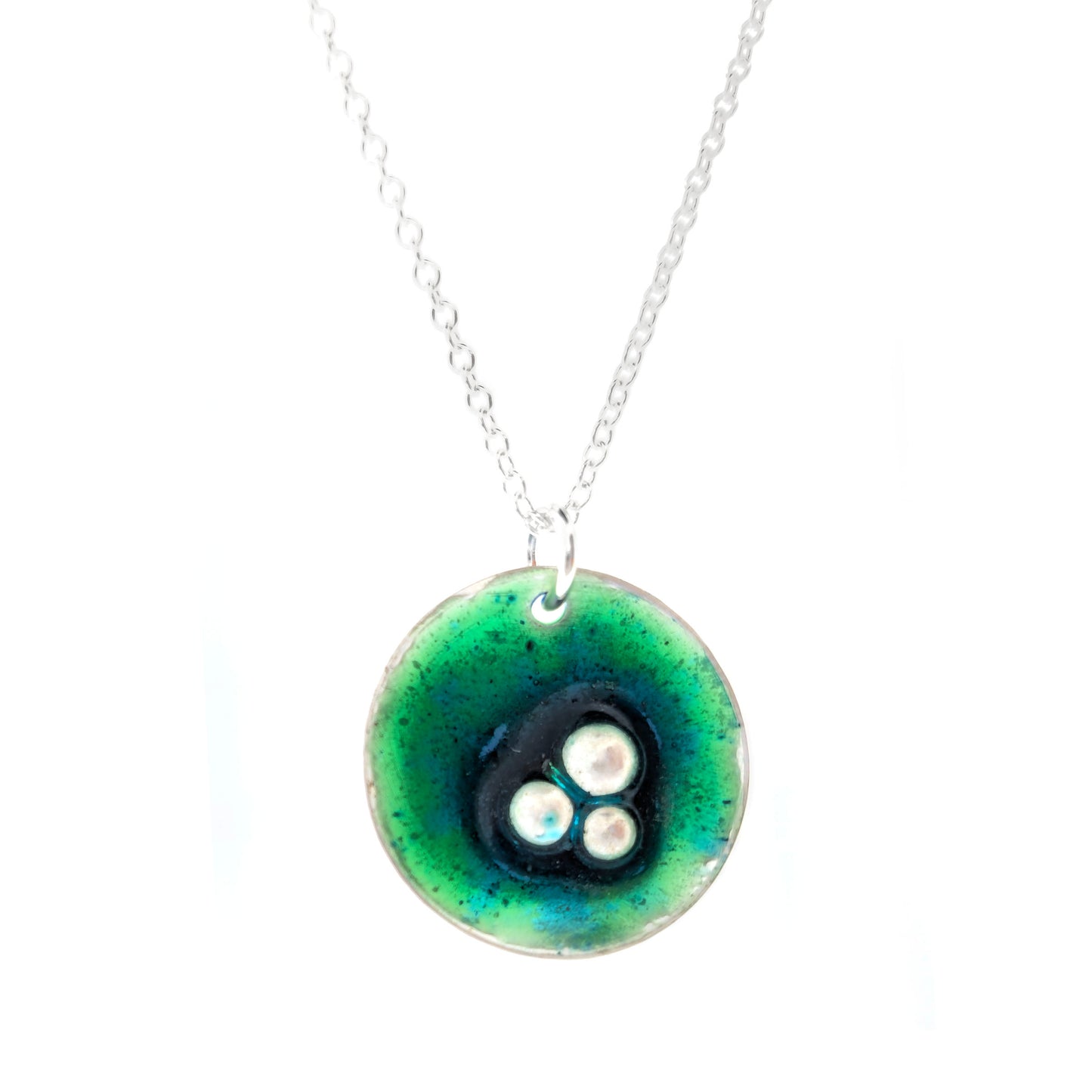 A handmade round silver pendant with a mix of green and turquoise enamel and three silver balls off-centre. Suspended on a silver chain. Shown on a white background. Mijoux Jewellery.