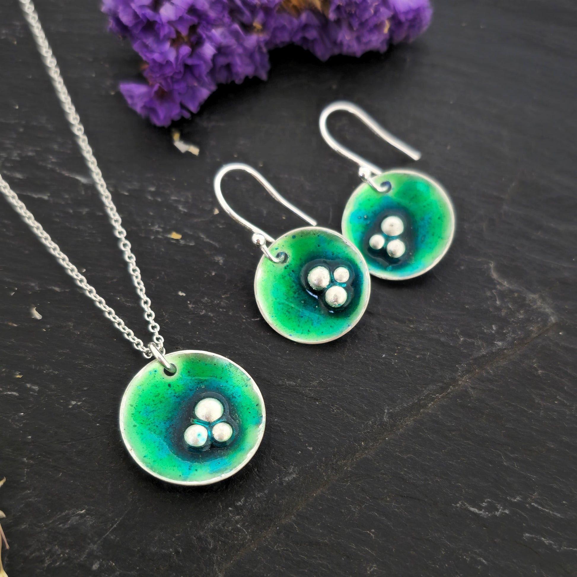 Handmade silver round drop earrings and pendant with green & turquoise enamel and 3 silver balls off centre. Shown on a black background with flowers. Mijoux Jewellery.