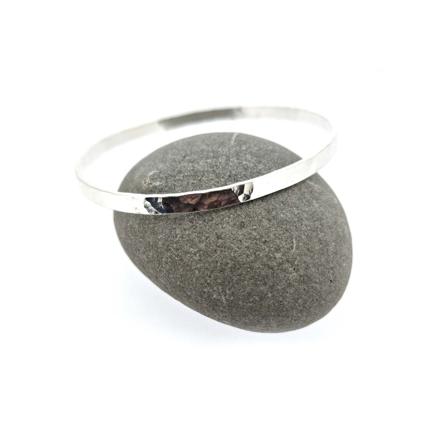 Silver round 4mm wide flat hammered bangle. Shown on pebble.